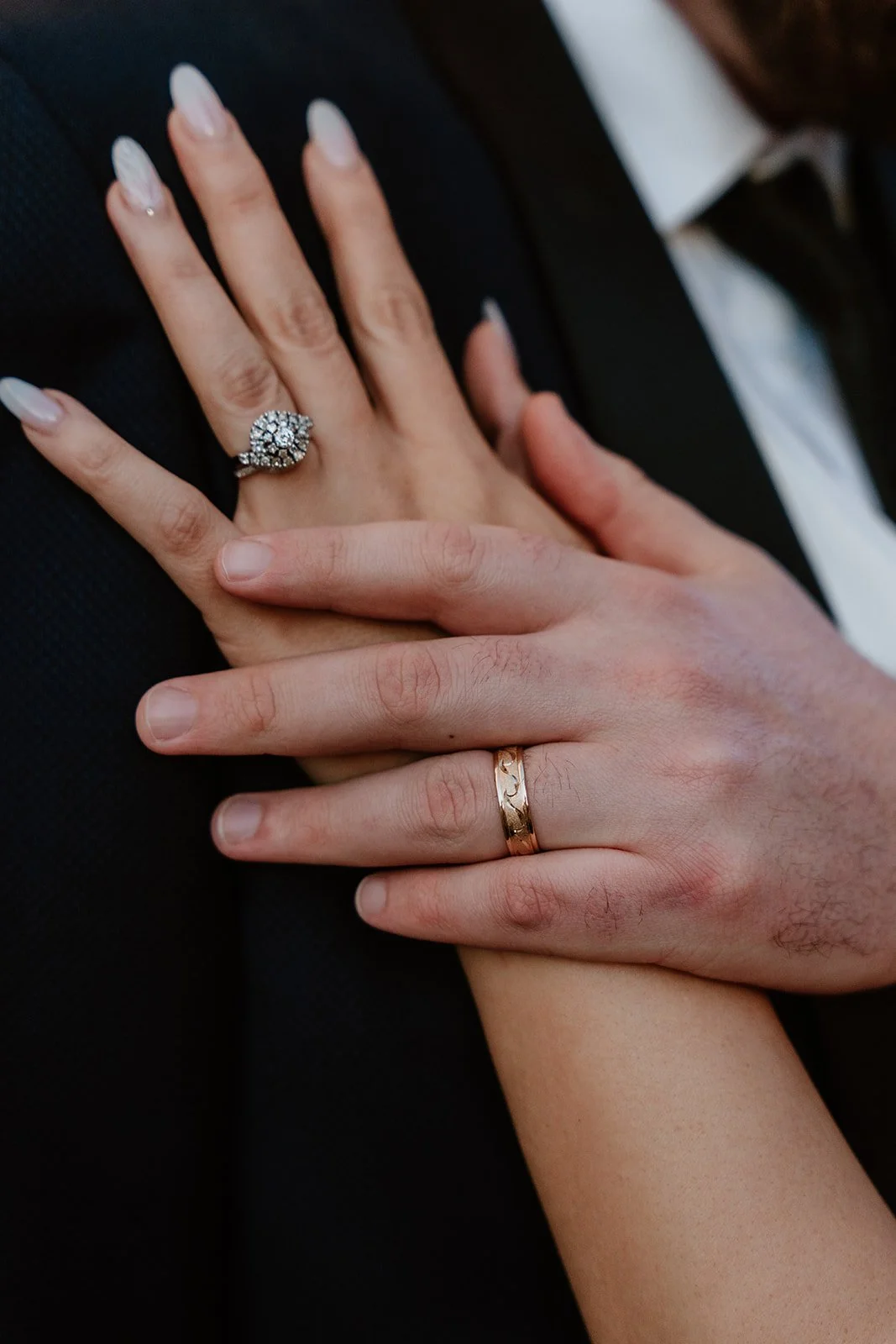 Close-up of a couple's hands showing wedding bands. The woman's hand has a large diamond engagement ring and a silver wedding band, while the man's hand has a gold wedding band. The woman has manicured nails with white tips and rhinestone accents.