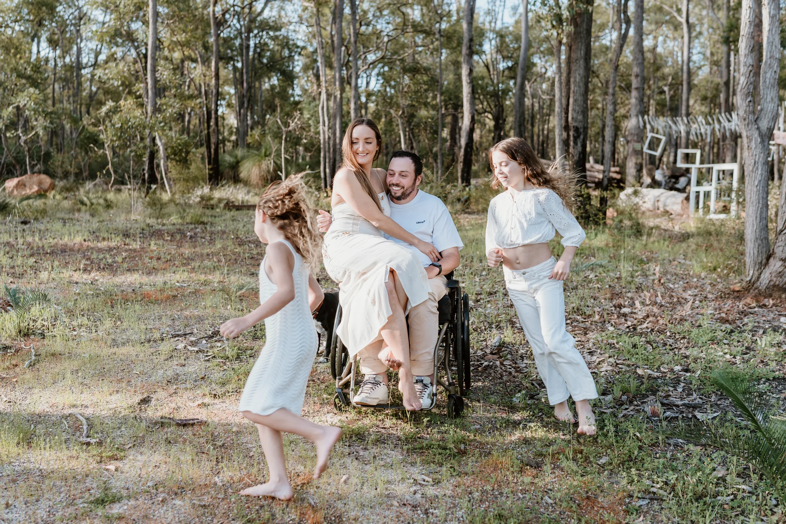 Family with children playing outdoors in a wooded area, some barefoot, engaging in playful activities, with trees and window frames hanging in the background.