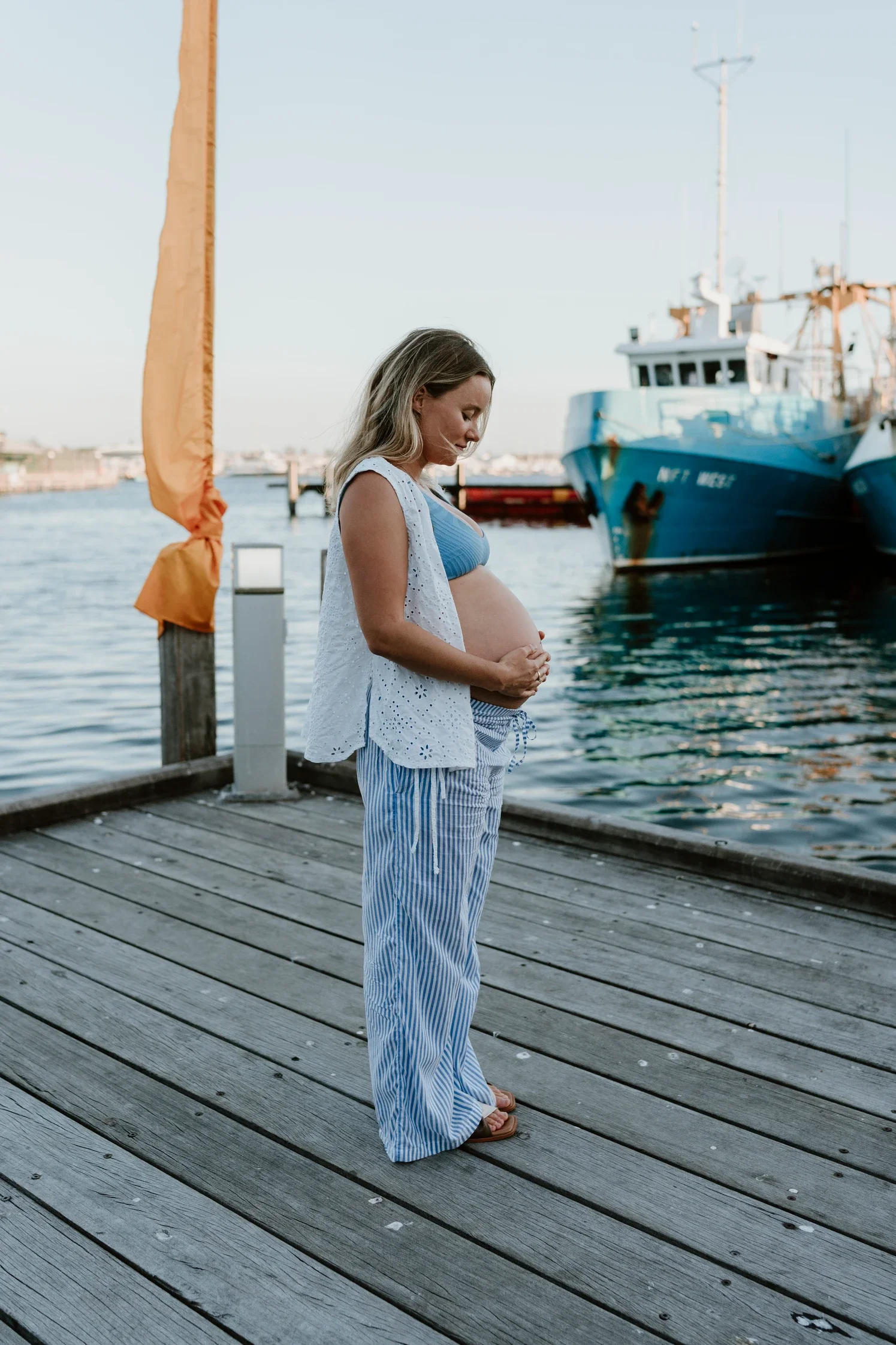 Pregnant woman standing on a wooden dock by the water, holding her belly, with boats docked in the background.
