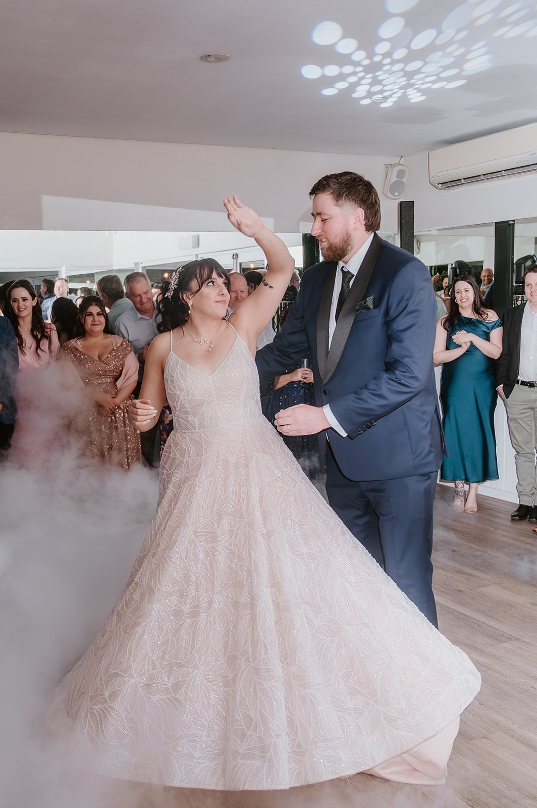 Bride and groom dancing at their wedding reception, with guests watching in the background, in a well-lit room with a wooden floor.