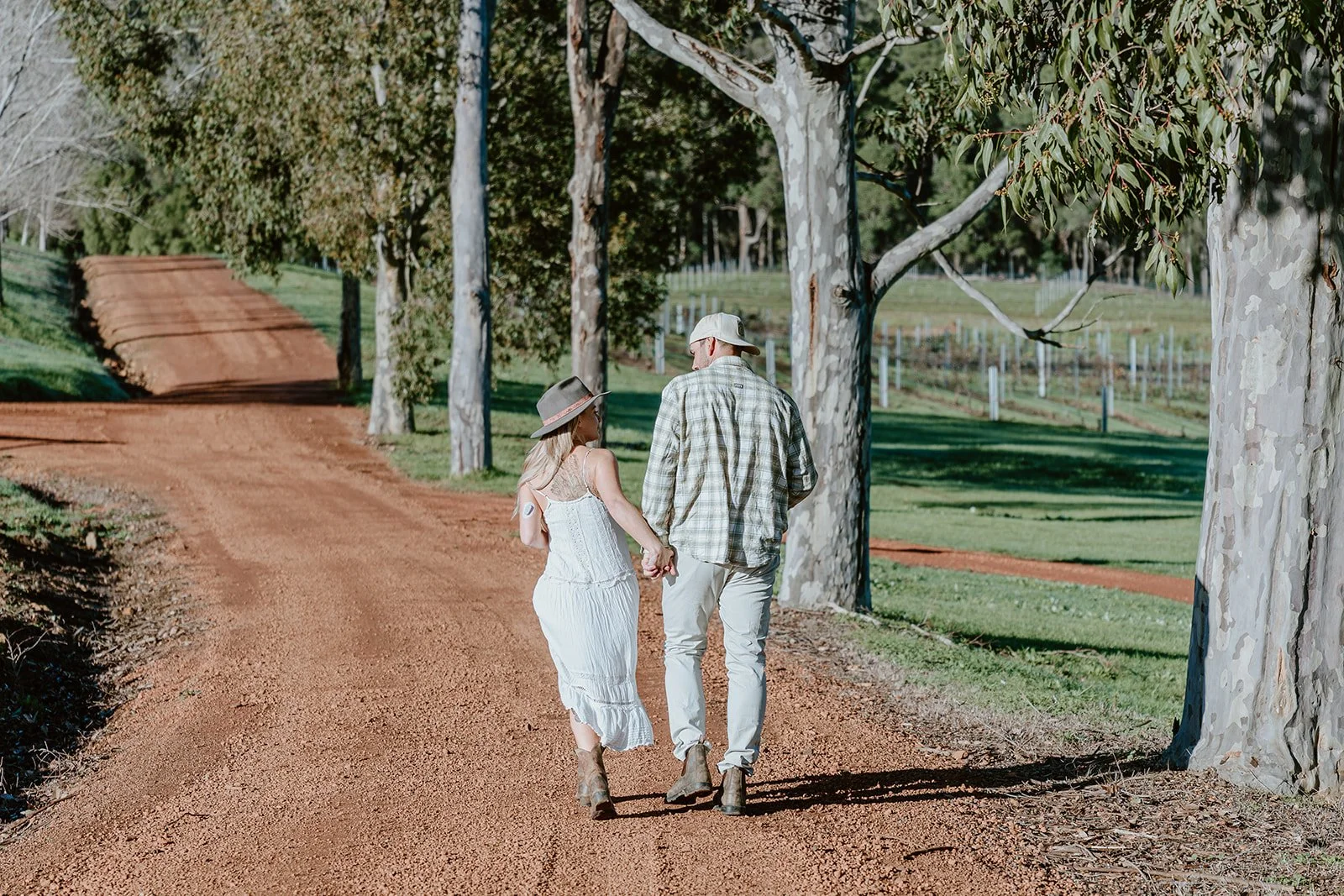 A couple walking hand in hand on a dirt road surrounded by tall eucalyptus trees in a rural area.
