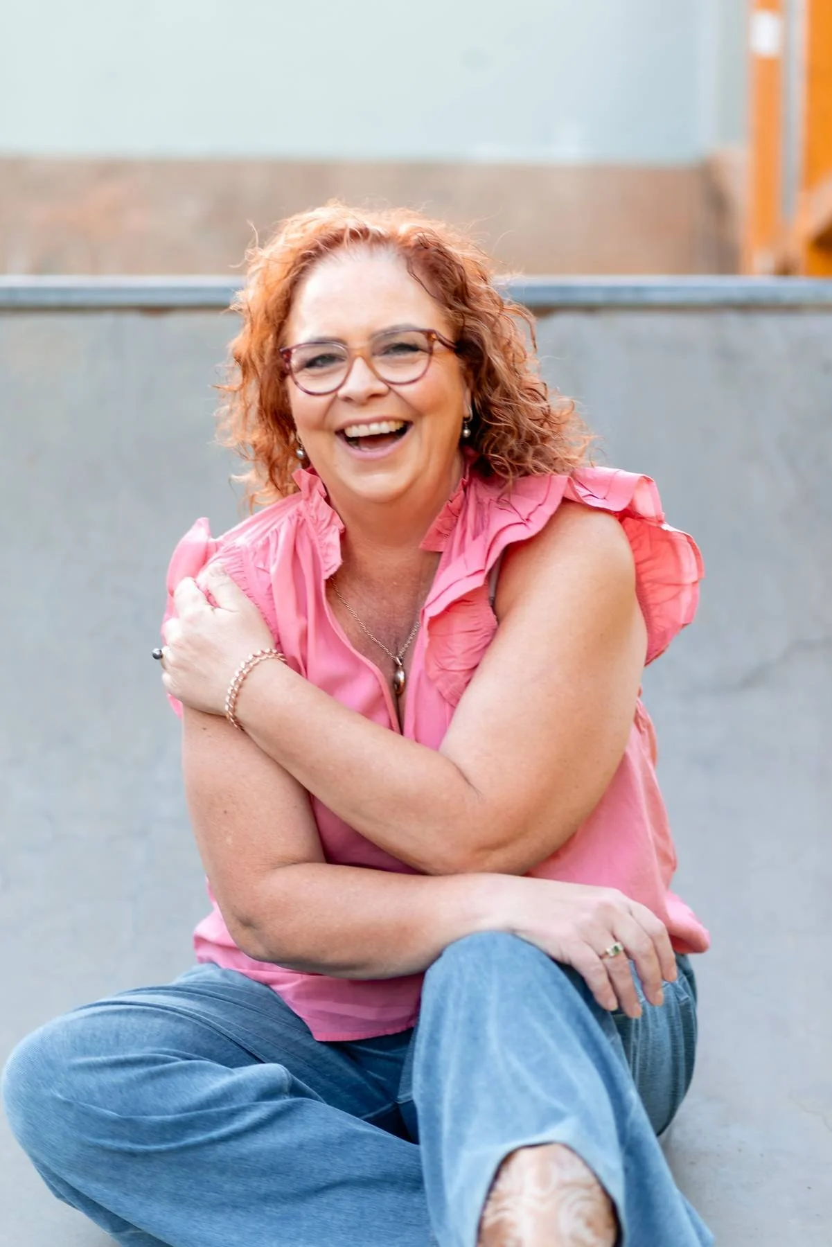 A woman with curly red hair and glasses, smiling and sitting outdoors, wearing a pink sleeveless top and blue jeans.