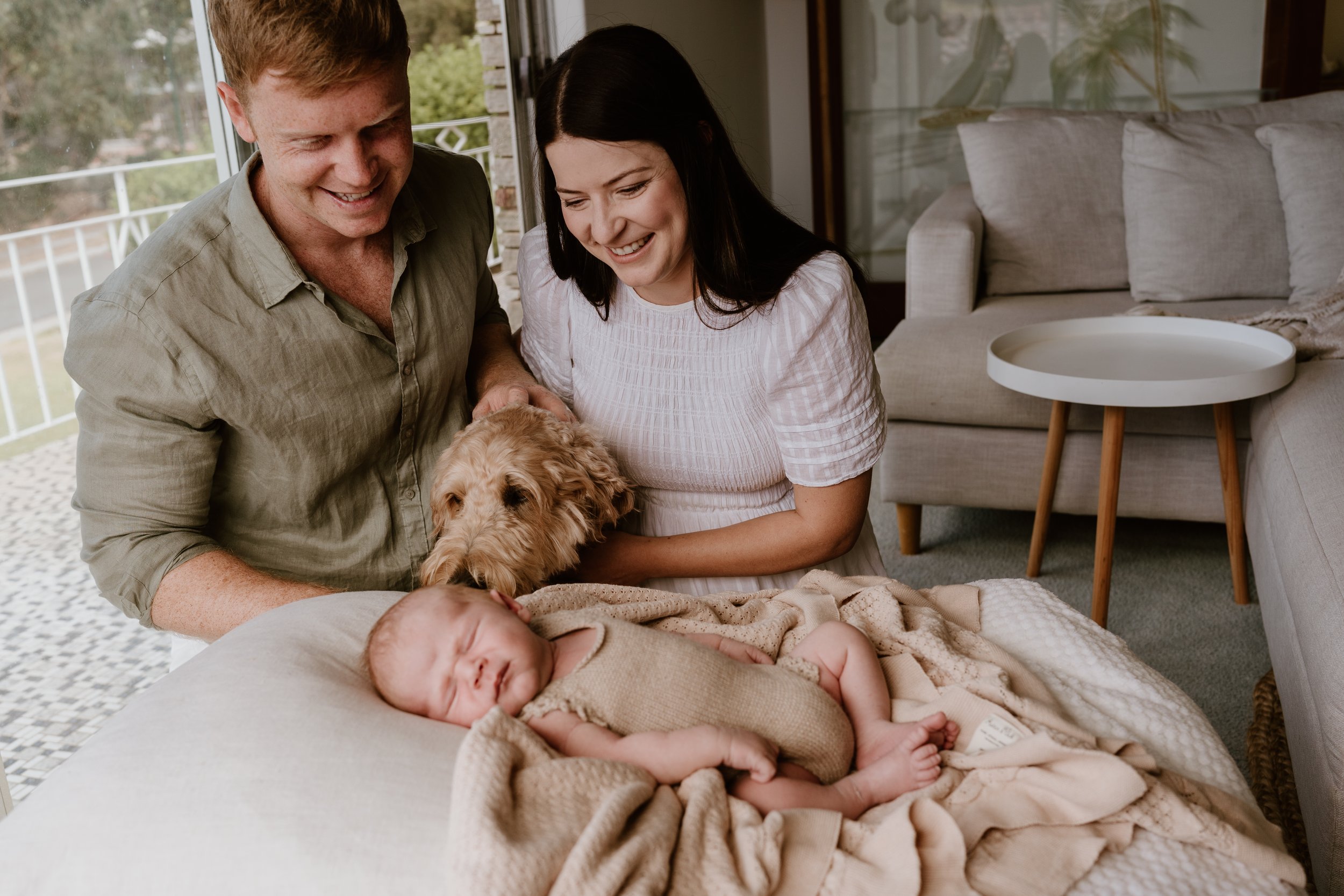 A happy family with two adults, a baby, and a dog gathered around the baby resting on a blanket.