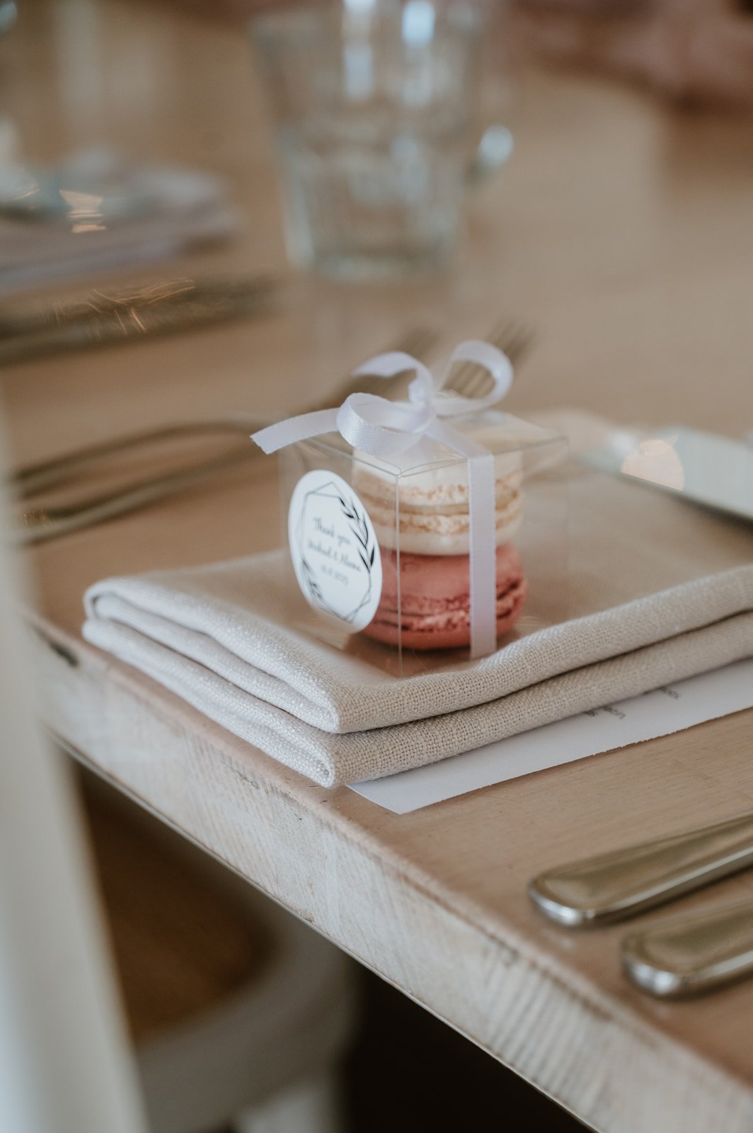 A small transparent box with two macarons, one pink and one cream, tied with a white ribbon, placed on a folded beige napkin on a wooden table. There are utensils and glassware in the background.