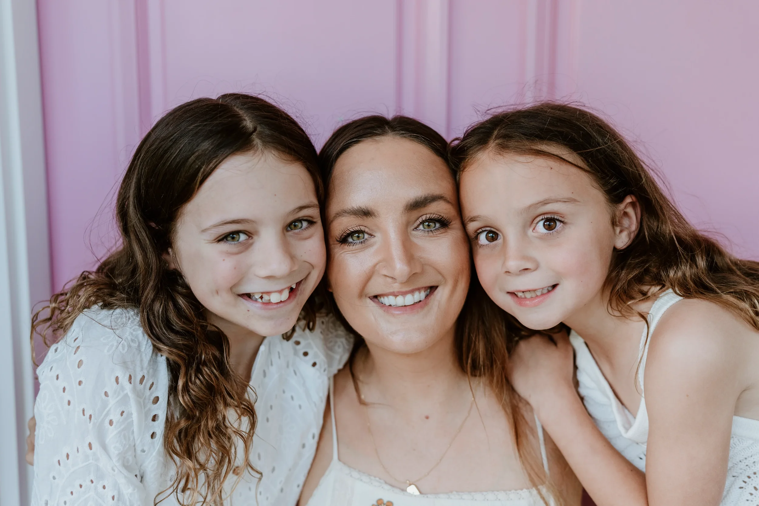 A smiling woman with two young girls standing close together against a pink background.