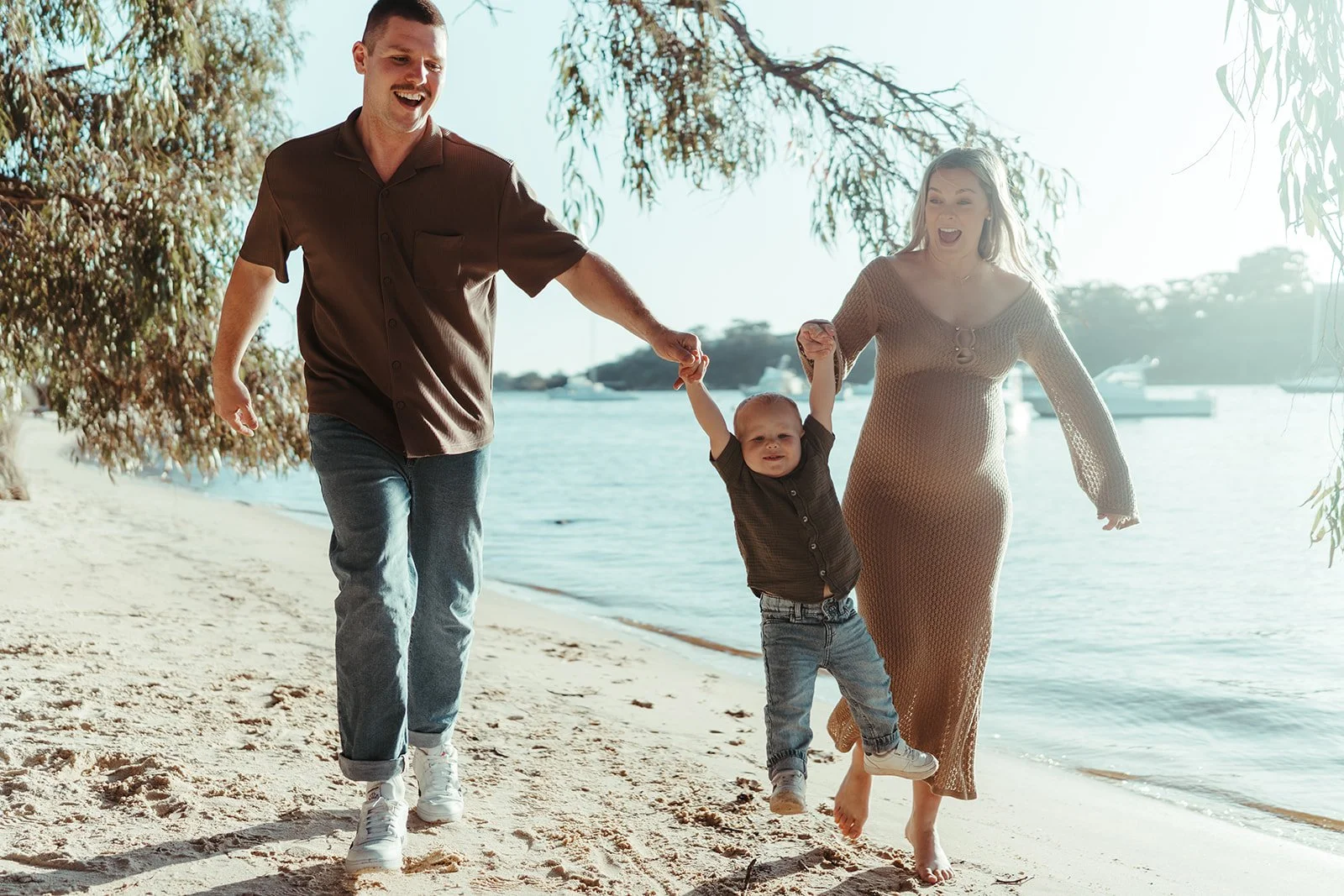 A family of three, a man, a woman, and a young boy, holding hands and walking barefoot on a sandy beach by the water with boats in the background.
