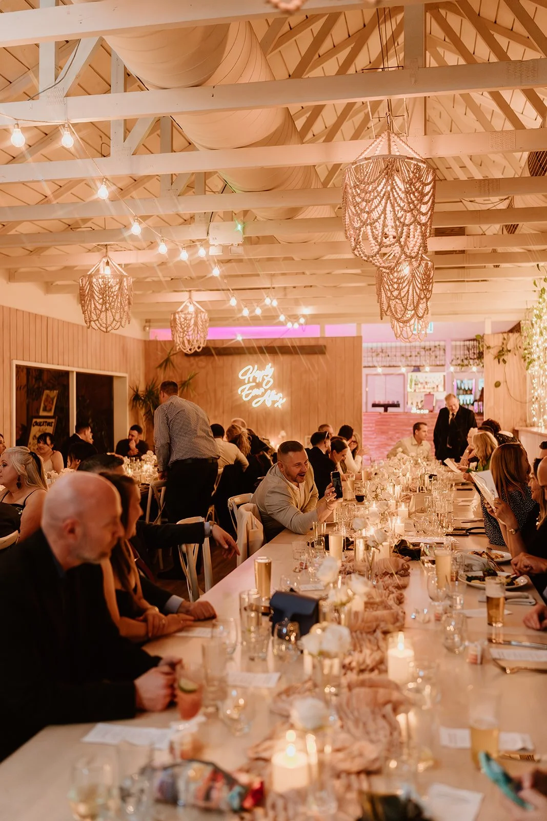 People seated at a long banquet table in a decorated event space with warm lighting and chandeliers, celebrating together.