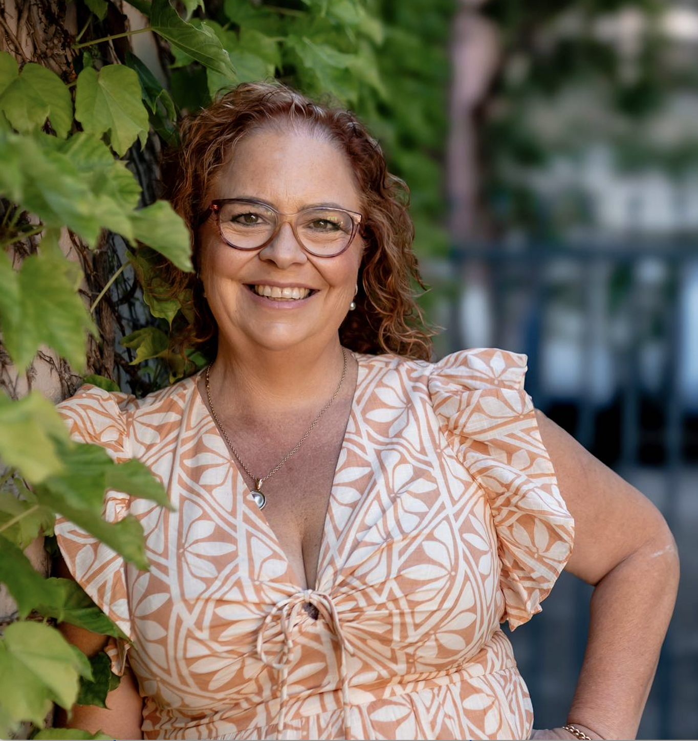 Smiling woman with curly red hair and glasses outdoors next to a vine-covered wall.