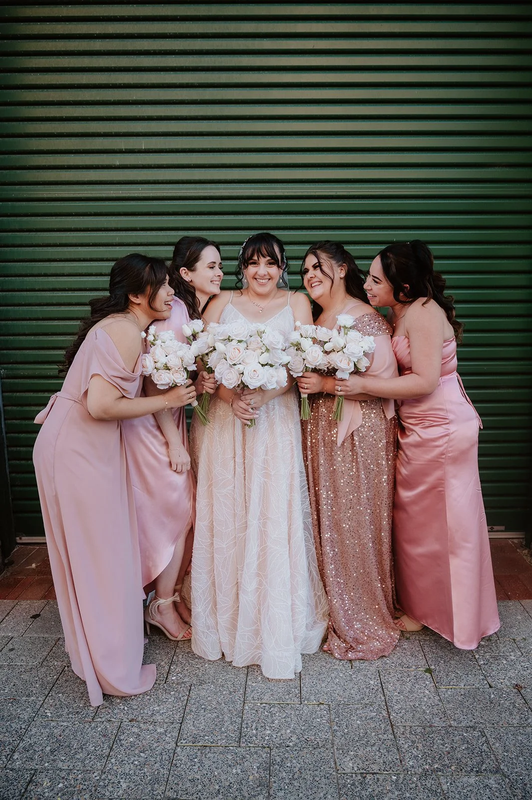 Group of five women, including a bride in a white wedding dress, holding white roses, standing in front of a green metal shutter, smiling and enjoying the moment.