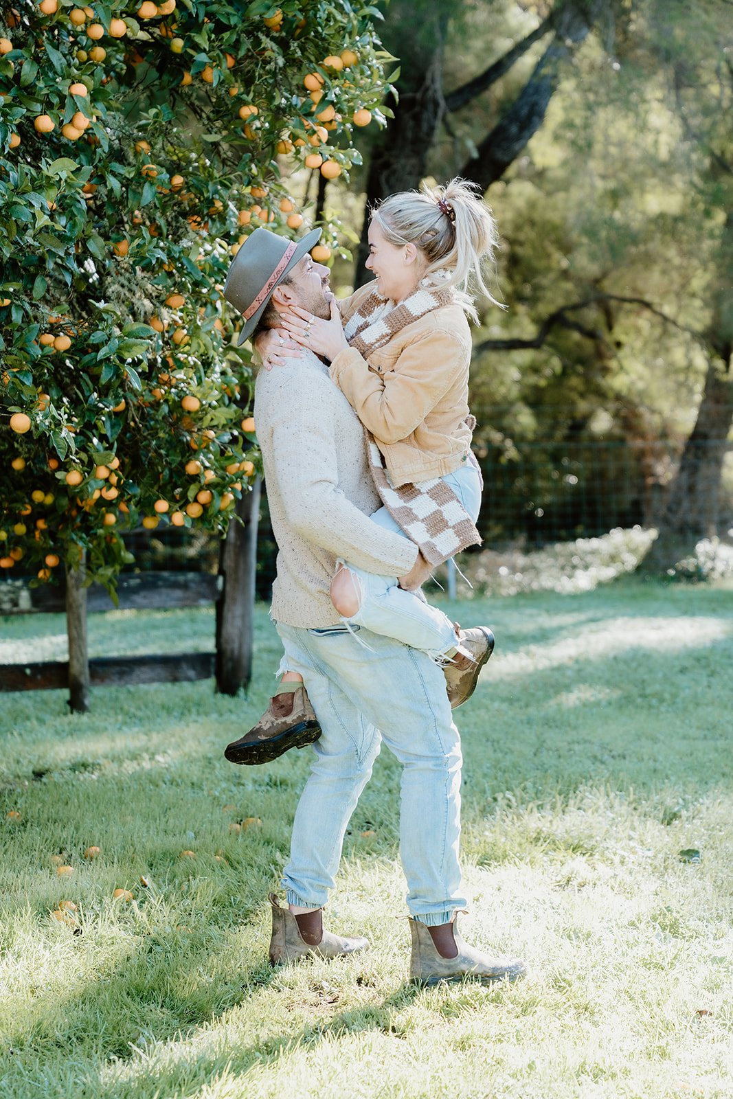 A man lifting a woman in front of an orange tree, both smiling and looking at each other, outdoors in a sunny, green park.