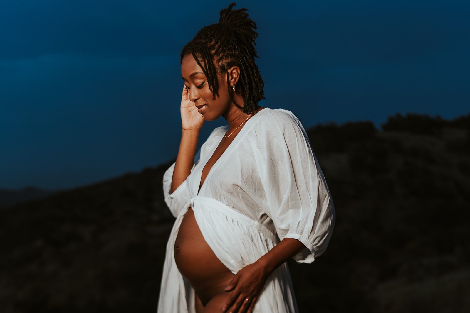 A pregnant woman with dark skin and dreadlocks, wearing a white dress, standing outdoors during evening or dusk, gently touching her belly and smiling softly.
