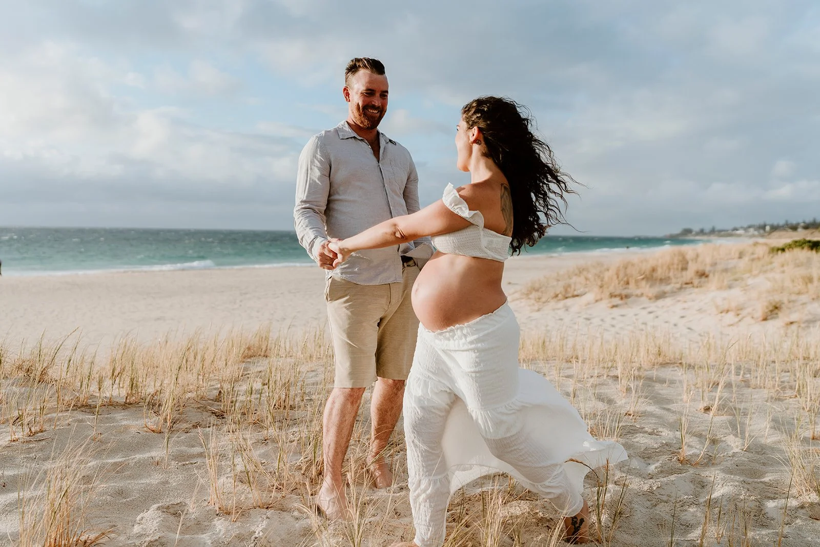 A pregnant woman and a man holding hands on a beach, smiling at each other, with the ocean and cloudy sky in the background.