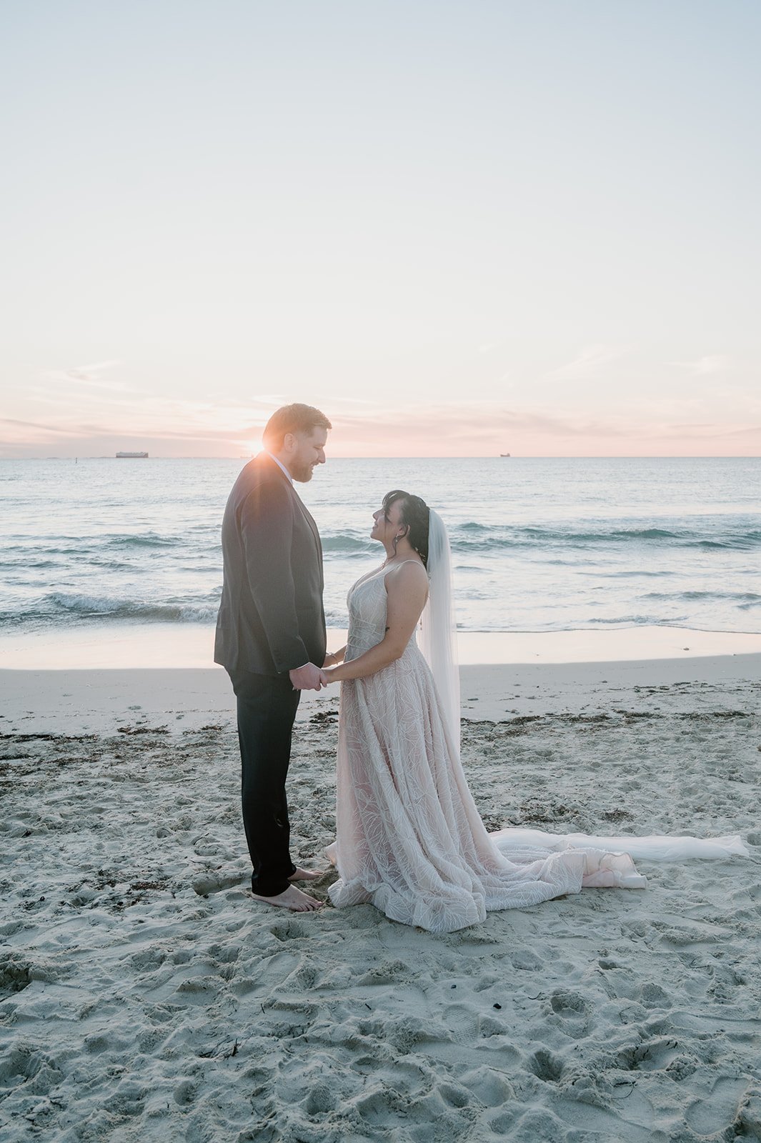 A bride and groom standing barefoot on the beach holding hands during sunset, with the ocean and sky in the background.