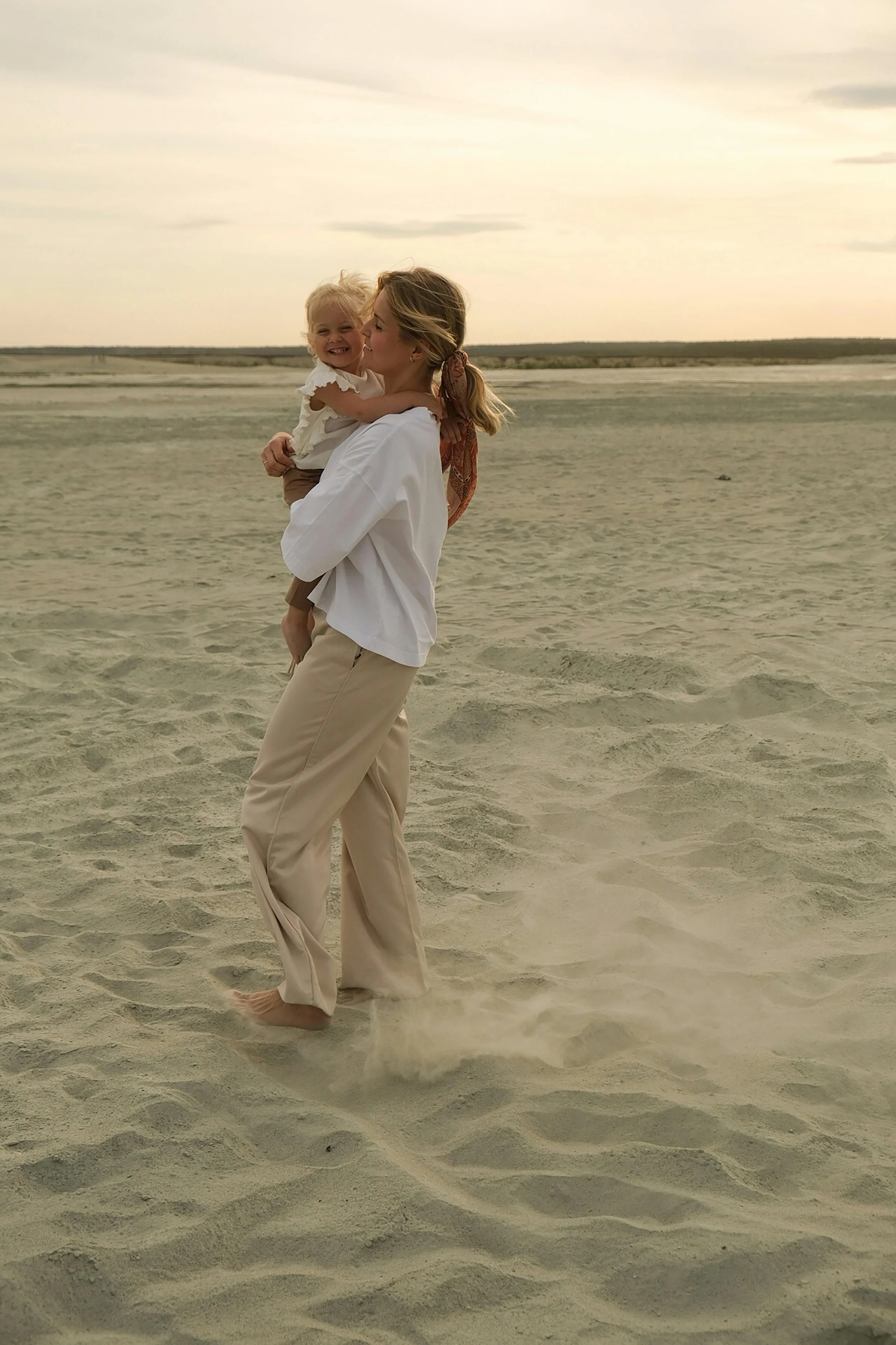 A woman lifting a smiling young girl on a sandy beach during sunset.