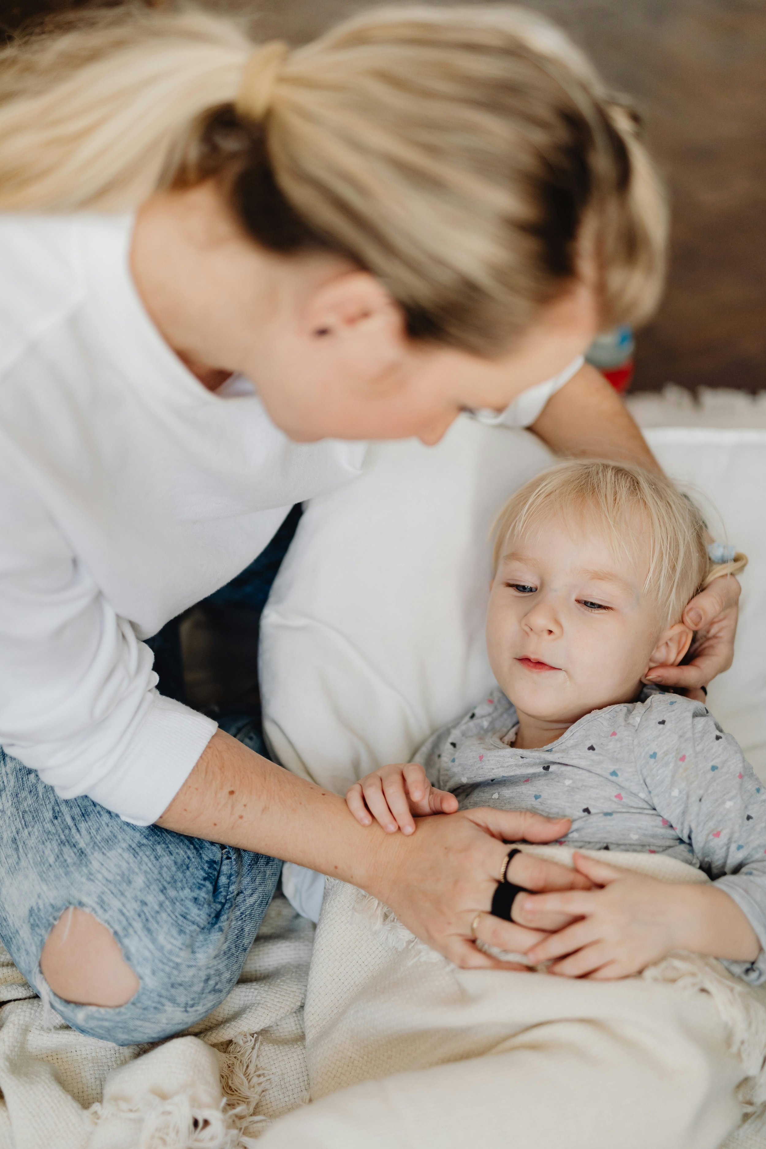 A woman with blond hair lies beside a young boy with blond hair, who is in bed. She is gently holding his arm and looking at him affectionately.