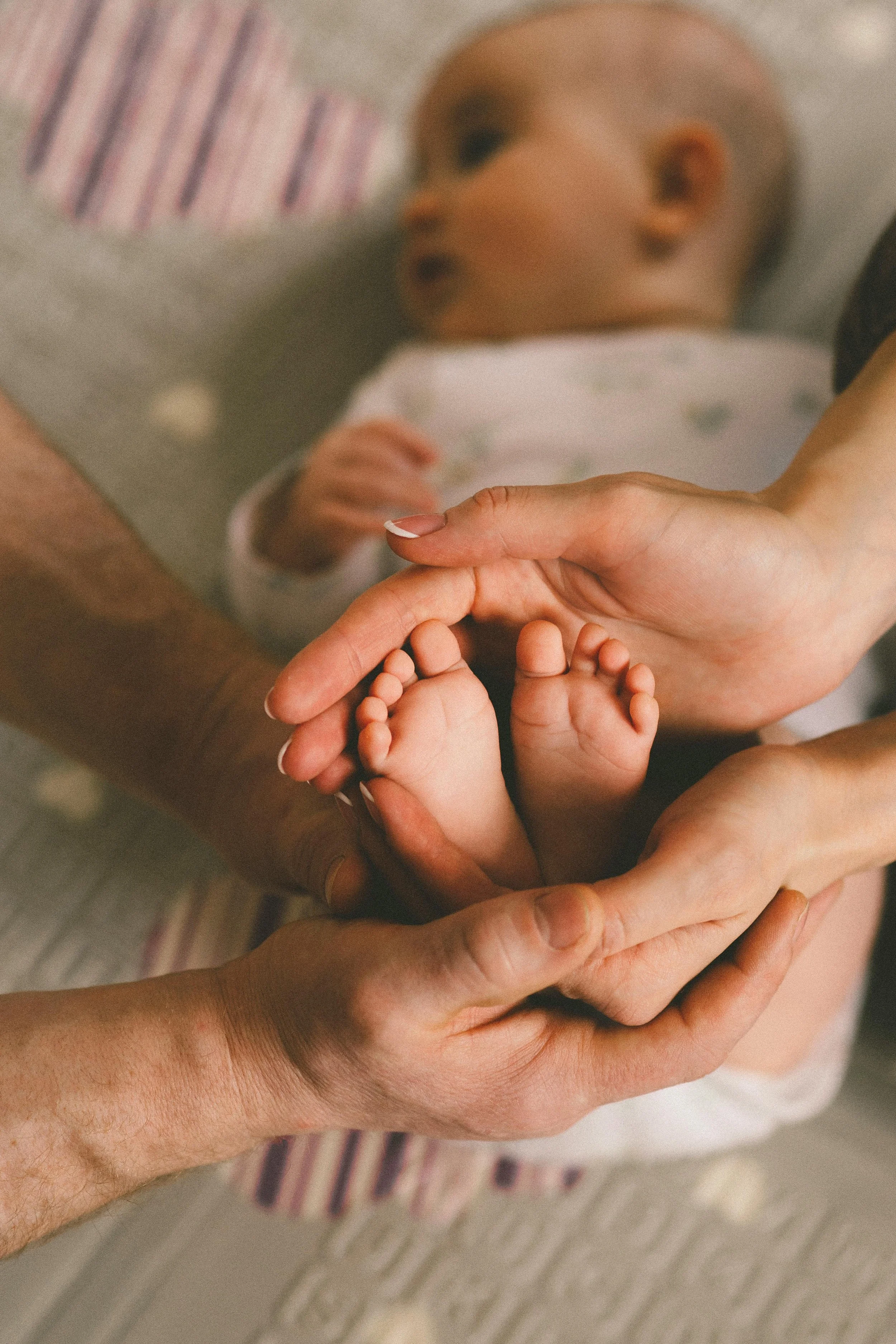 Close-up of adult hands gently holding tiny baby feet, with a blurred infant lying in the background.