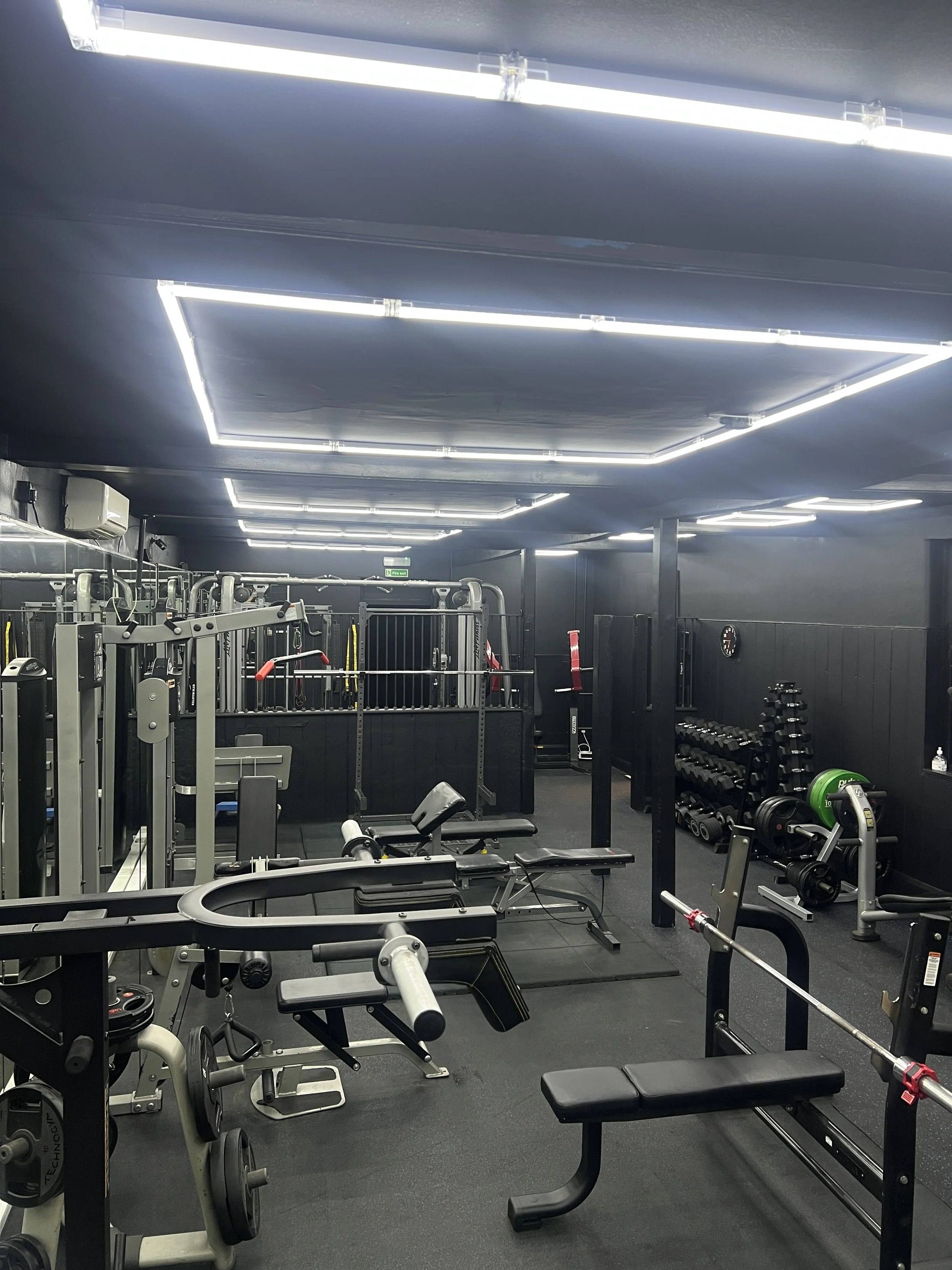 Interior of a gym with various workout equipment including weight machines, benches, dumbbells, and barbells, with black walls and ceiling illuminated by bright white LED lights.