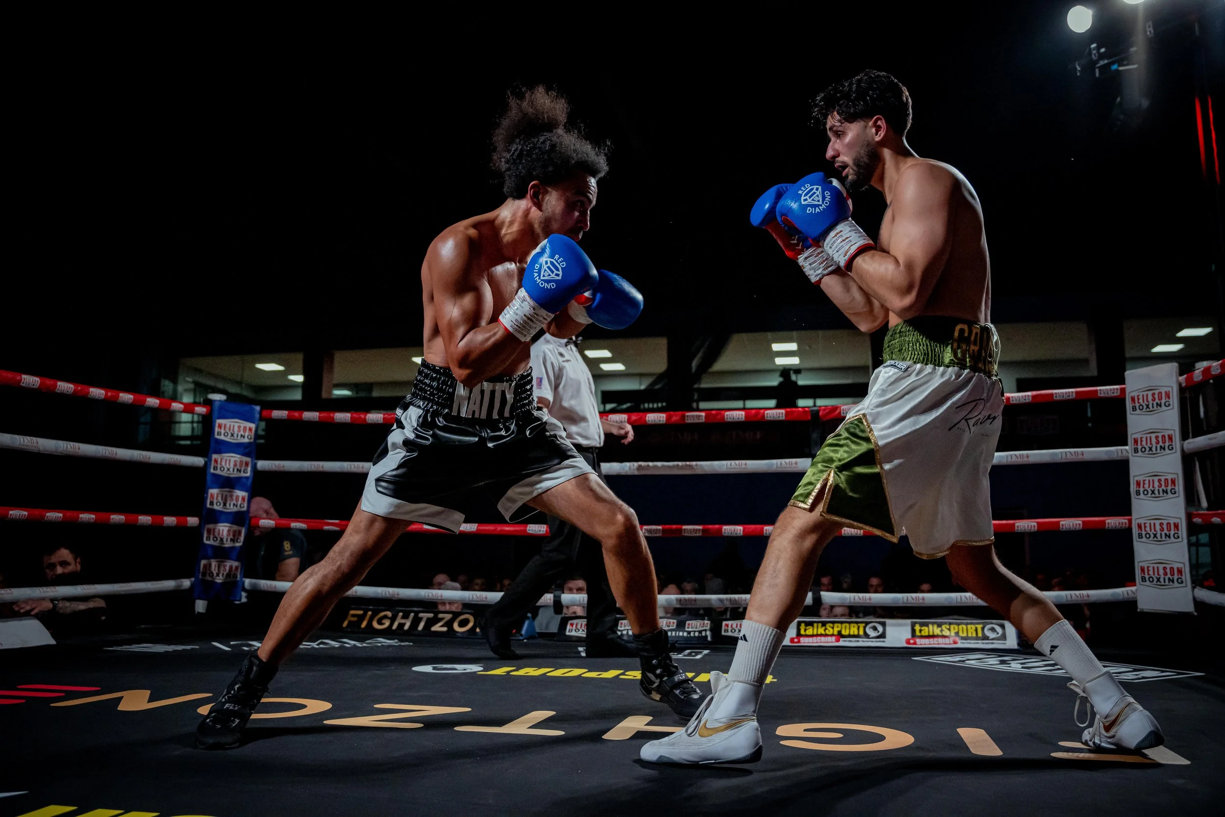 Two male boxers in a ring, facing each other in a boxing match, wearing boxing gloves and shorts, with a referee in the background, under focused lighting.