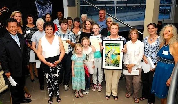Group of people, including children and adults, gathered indoors for a photo, some holding certificates and an art framed picture, in a well-lit room with large windows.