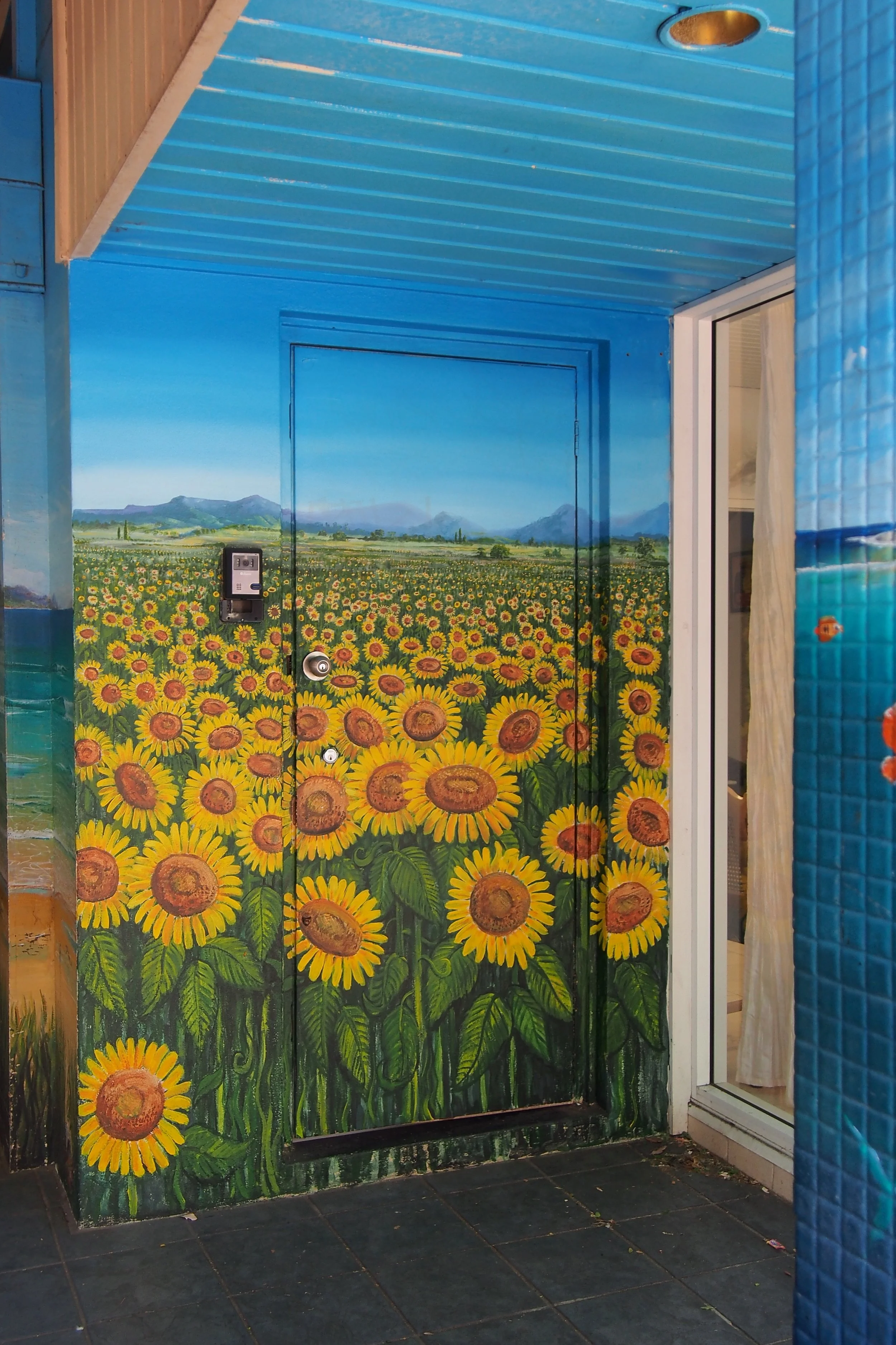 Door and wall with a sunflower field painting, with blue sky and mountains in the background, and a sliding glass door to the right.