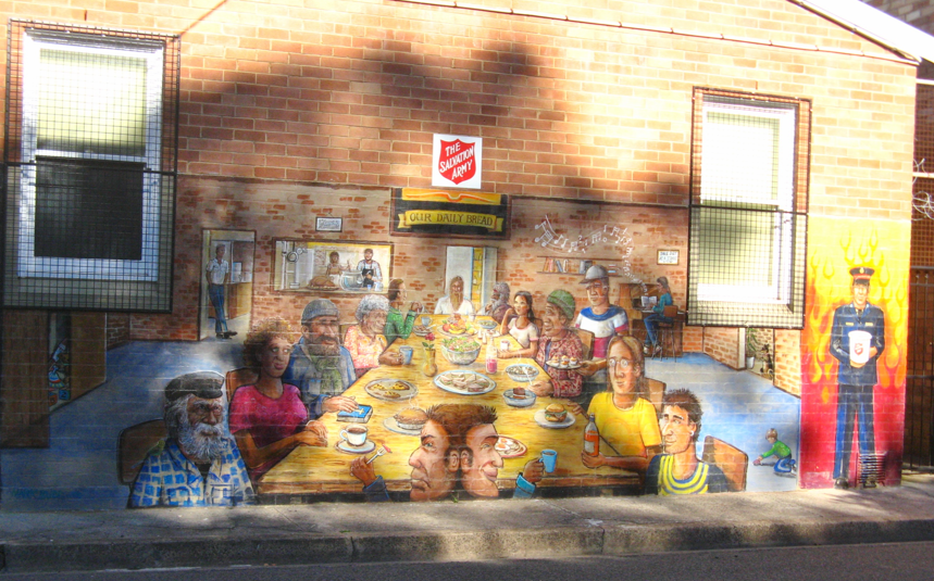 Colorful mural on a brick wall depicts a diverse group of people sitting around a dining table with breakfast food, in front of a building with large windows and various signs, including 'The Salvation Army' and 'Our Daily Bread'.