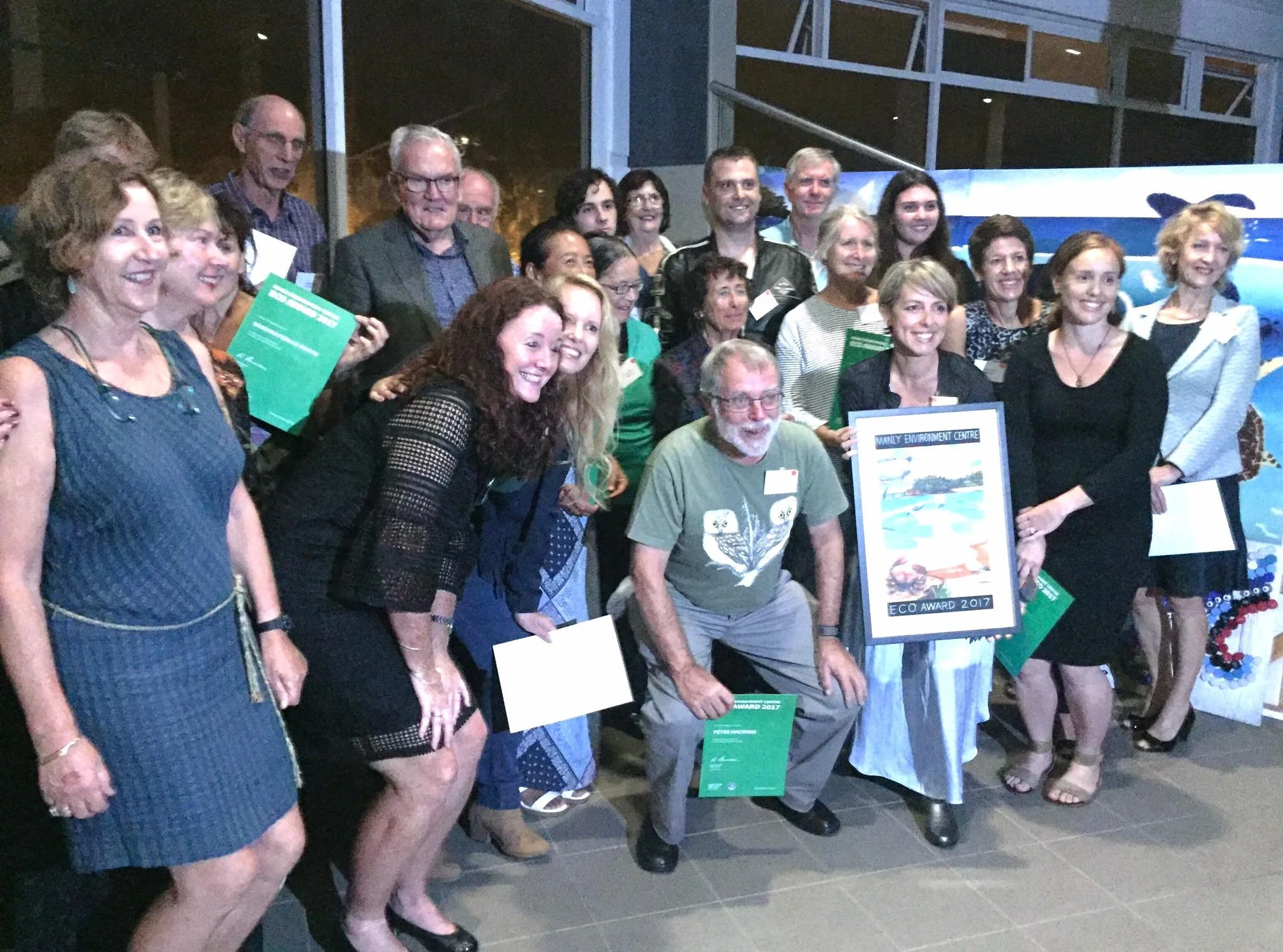 Group of people at an award ceremony, some holding certificates, gathered indoors with a large window in the background.