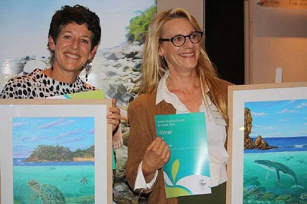 Two women smiling and holding framed artwork and certificates, standing indoors with a nature-themed backdrop.