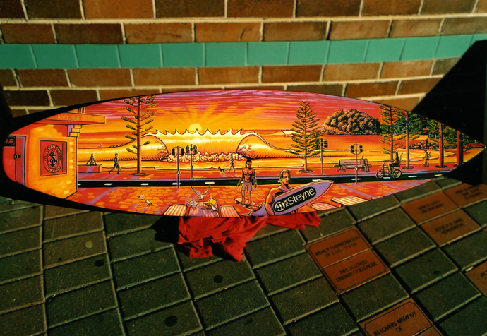 Colorful skateboard deck featuring a sunset scene with people, trees, and the ocean, placed on a brick ledge.