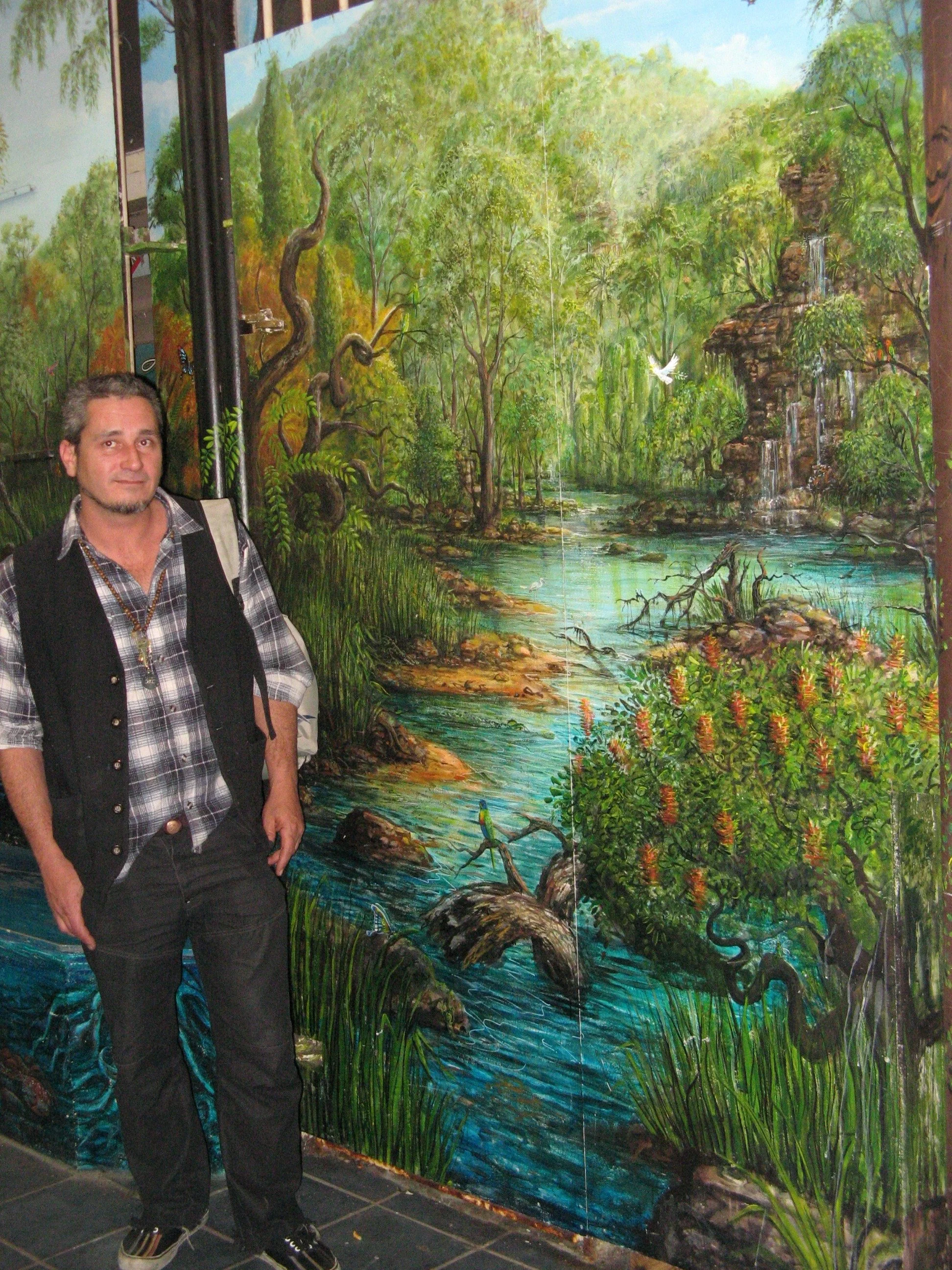 A man standing in front of a detailed landscape mural with trees, water, and wildlife.