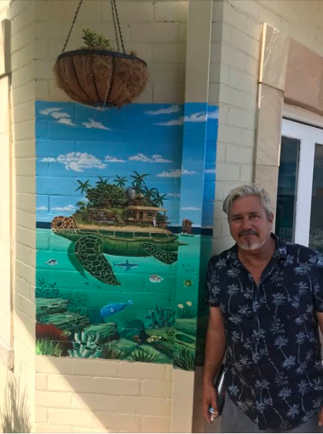 A man standing next to a colorful mural of an underwater scene with a turtle, coral, fish, and tropical plants painted on a brick wall, with a hanging basket above.