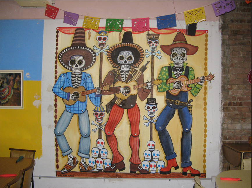 Colorful mural of three skeleton musicians in Mexican mariachi style, wearing sombreros and suits, surrounded by decorated skulls and papel picado banners.
