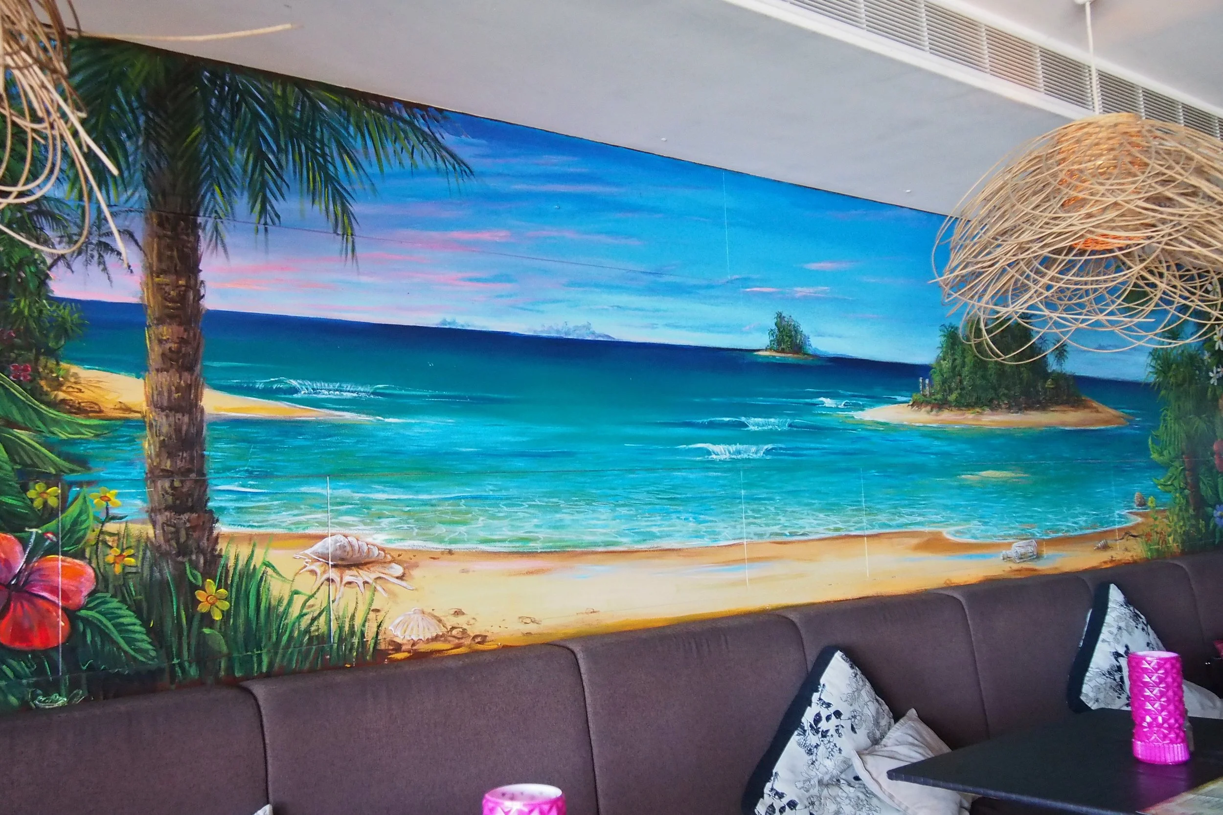 A vibrant mural depicting a tropical beach scene with turquoise sea, sandy shores, palm trees, and small islands under a pink and blue sky, located on a wall behind a dark-brown cushioned seating area.