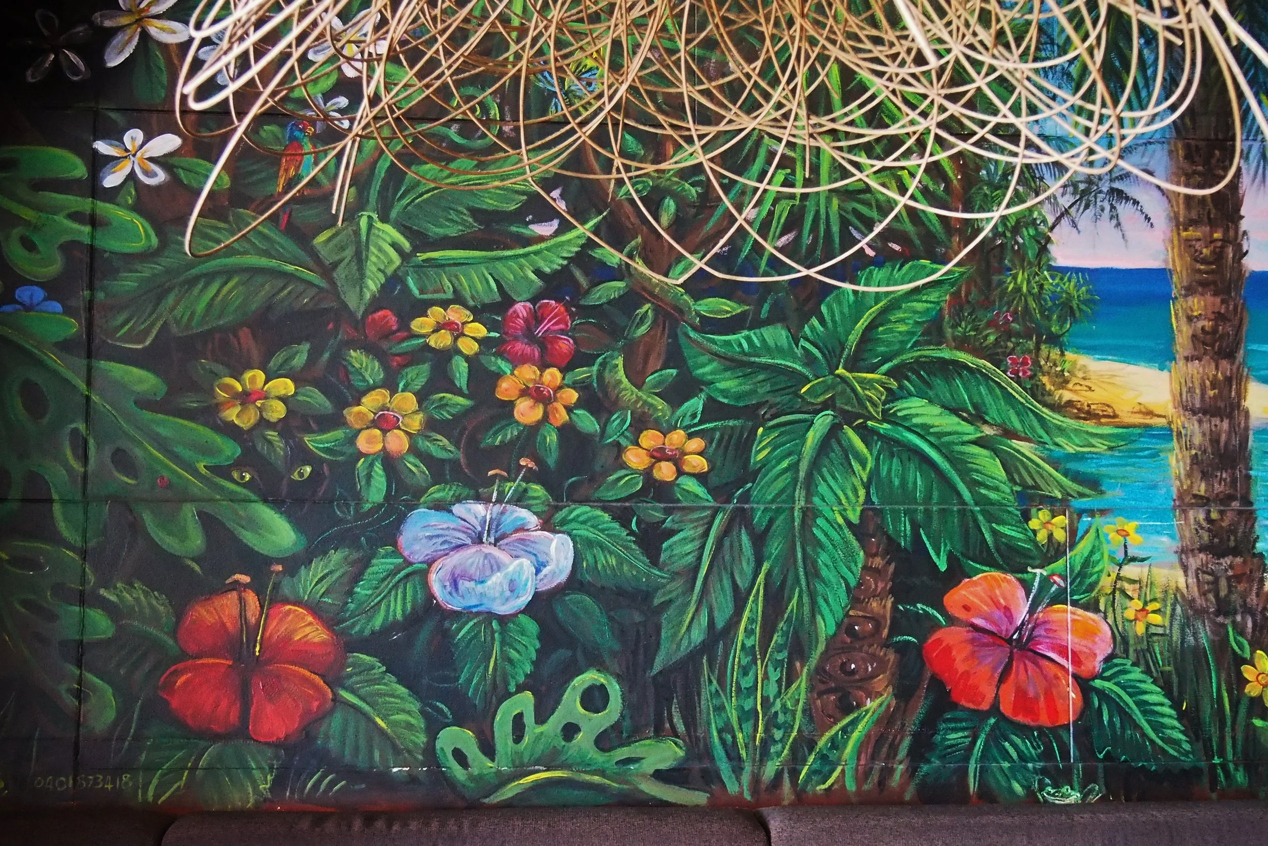 Colorful tropical mural featuring lush green leaves, vibrant flowers, a parrot, a cat hidden behind foliage, and a beach scene with the ocean in the background. There are string lights hanging at the top.