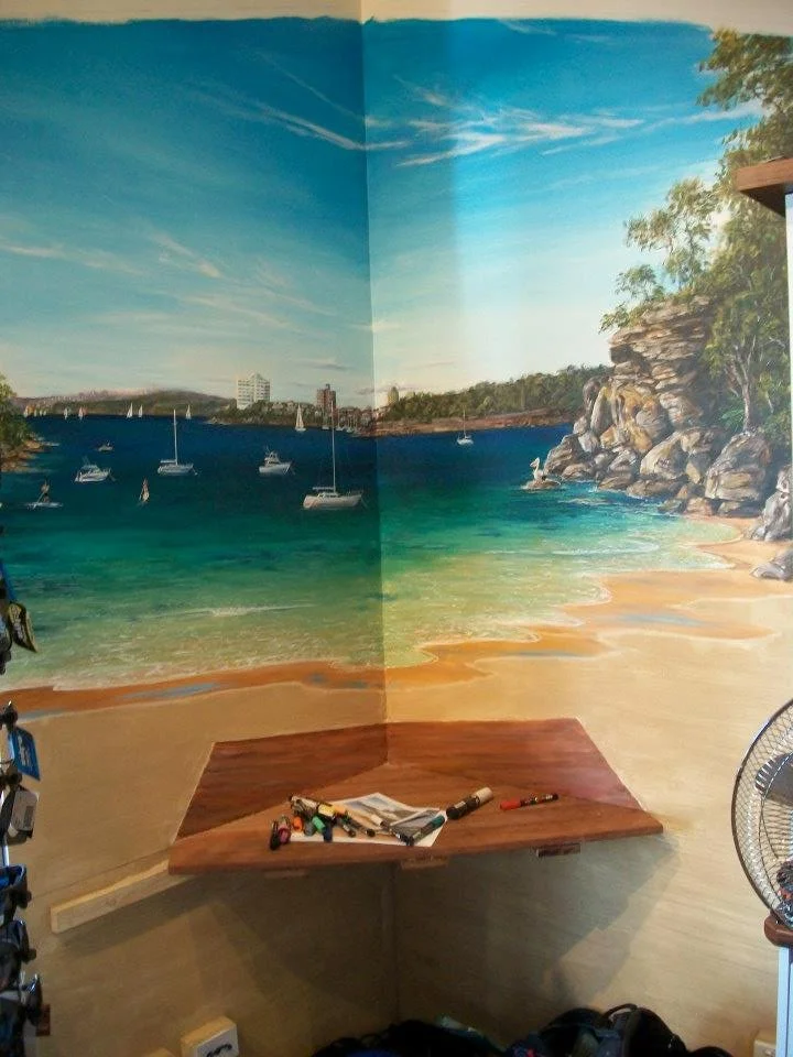 Colorful mural of a beach scene with blue sky, sailboats, and rocky cliffs on a wall in a room.