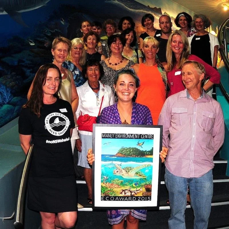 A group of people posing together, holding a colorful poster of a sea turtle and ocean scene at the Manly Environment Centre, with some individuals wearing name tags.
