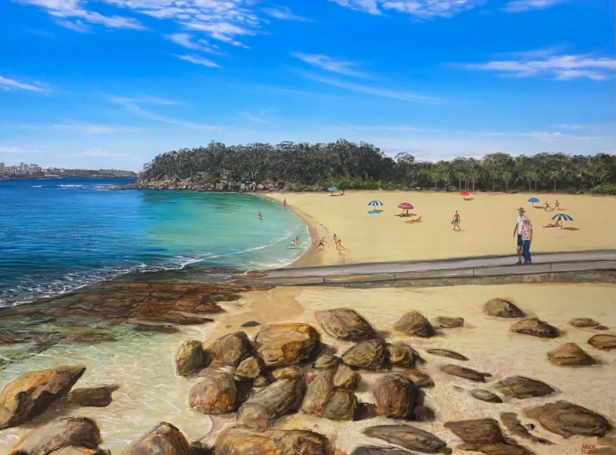 A sunny beach scene with people relaxing under umbrellas, swimming, and walking along the sandy shore, with rocky area in the foreground and a tree-covered hill in the background.