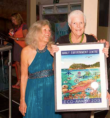 Two elderly women smiling and holding a framed award for the Manly Environment Centre, given in 2013, with a colorful aquatic scene in the background.