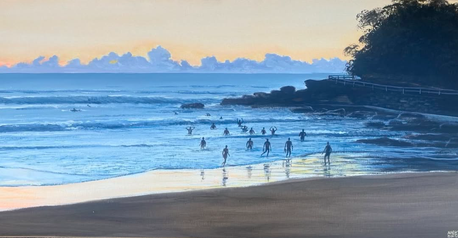 People are swimming and playing in the ocean waves at sunset on a beach, with a rocky shoreline and trees on the right.