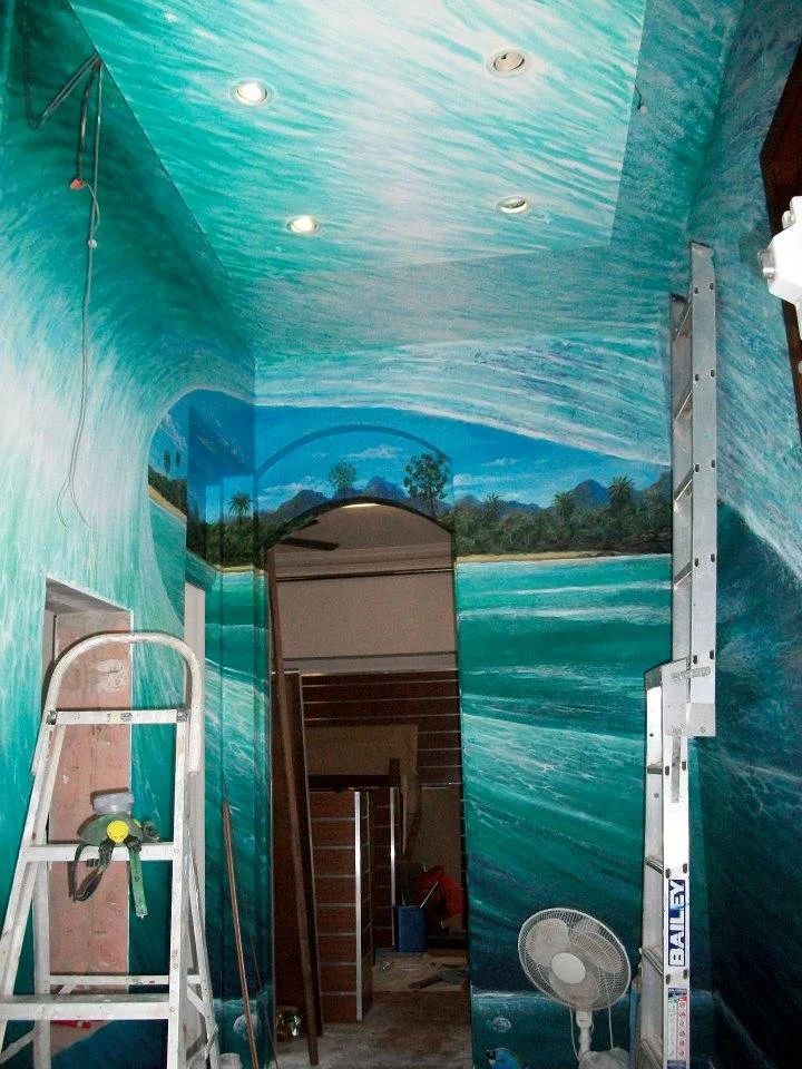 Interior of a room with walls and ceiling decorated with a mural of lakes and trees, featuring a mirror, a ladder, and a standing fan.