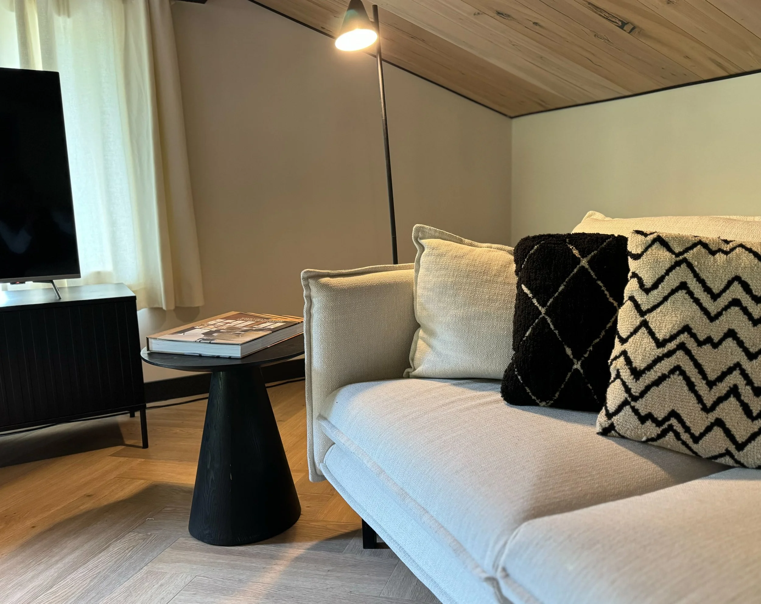 Living room with a white sofa, black and white throw pillows, a black side table with a magazine, a black floor lamp, a window with beige curtains, a wall with wood paneling ceiling, and hardwood flooring.