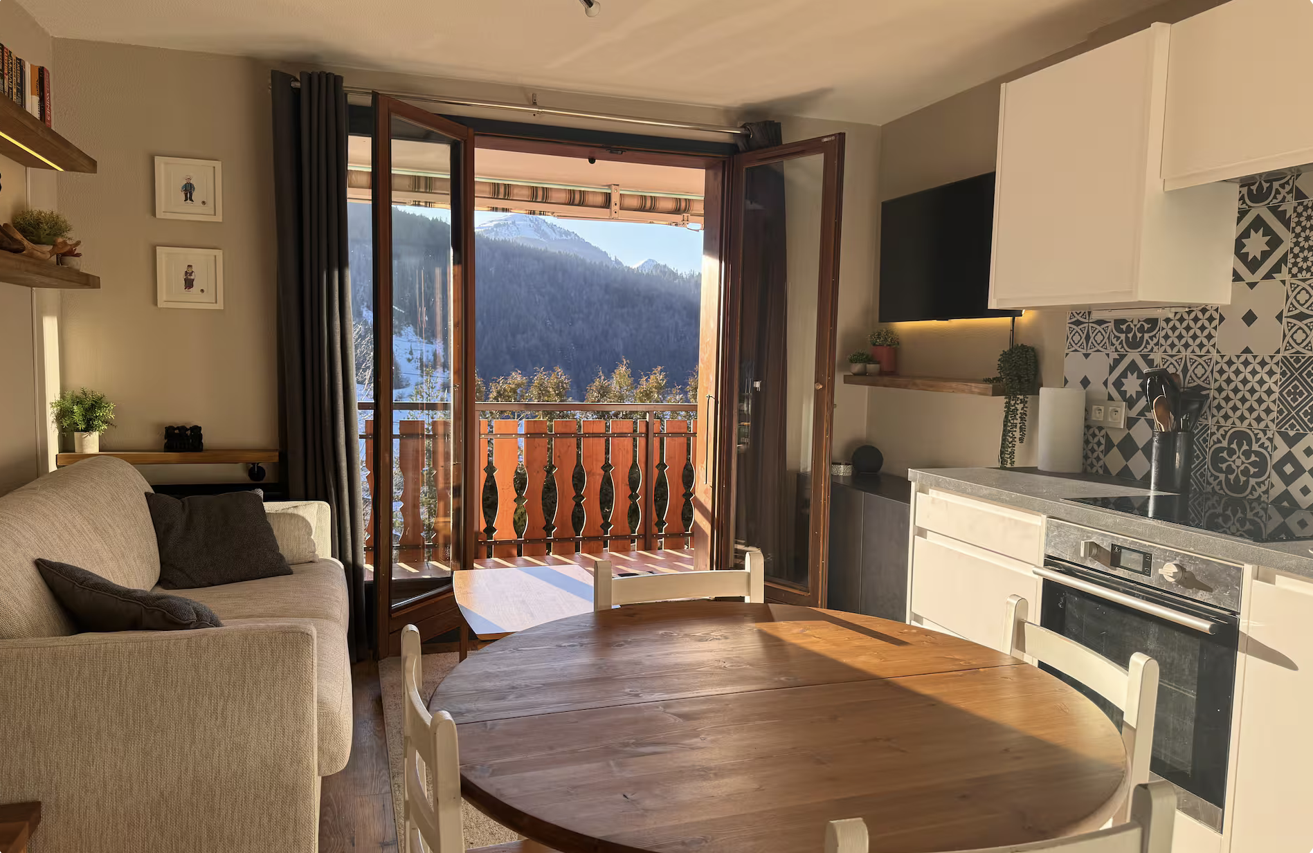 View from inside a cozy living room with a beige sofa, a round wooden dining table, and an open door leading to a balcony with mountain scenery in the background. The room features a patterned tile backsplash in the kitchen area and small decorative plants.