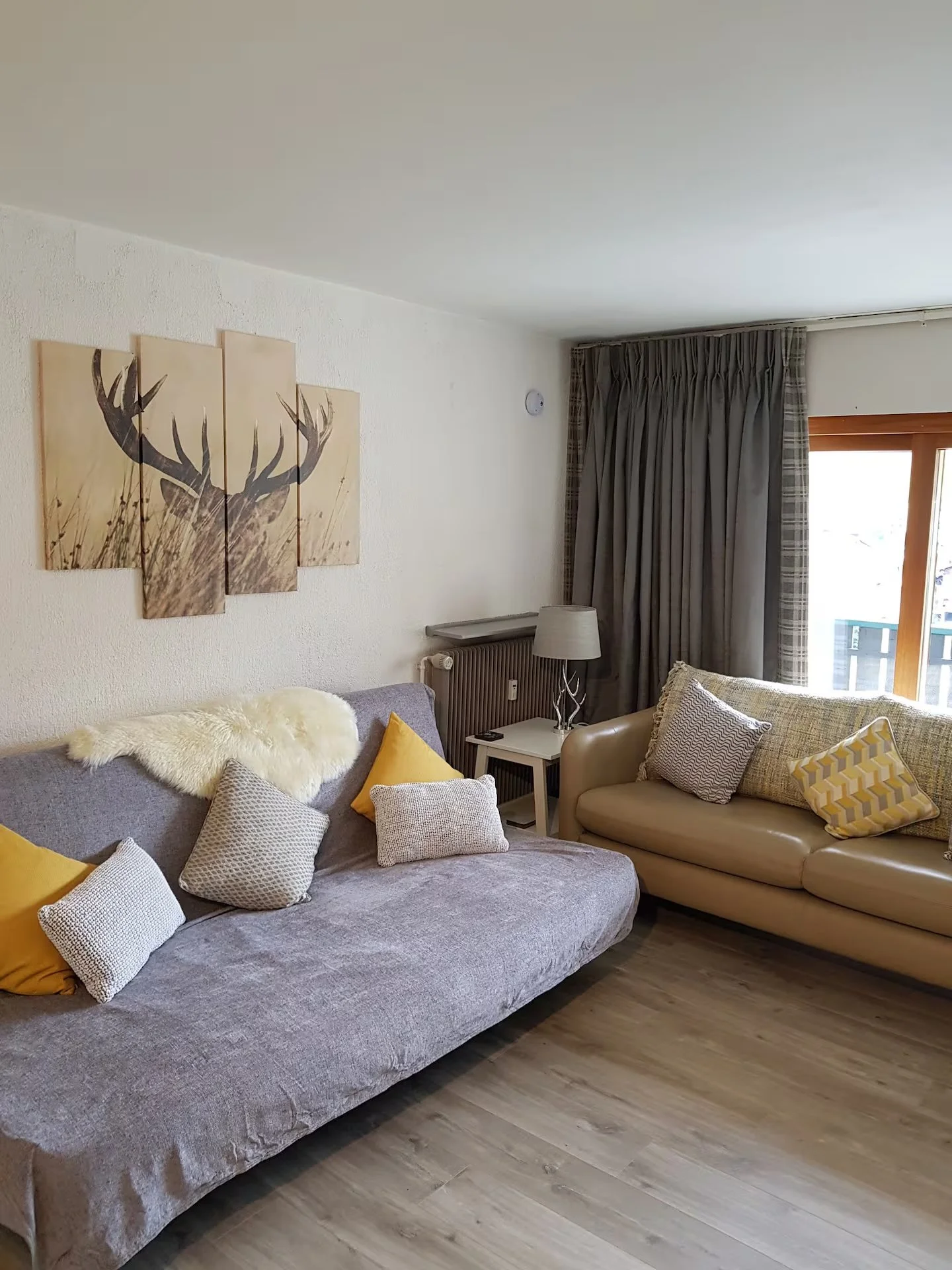 A living room with two sofas, multiple cushions, a small side table, a lamp, a window with curtains, a wall painting of a deer, and wood flooring.