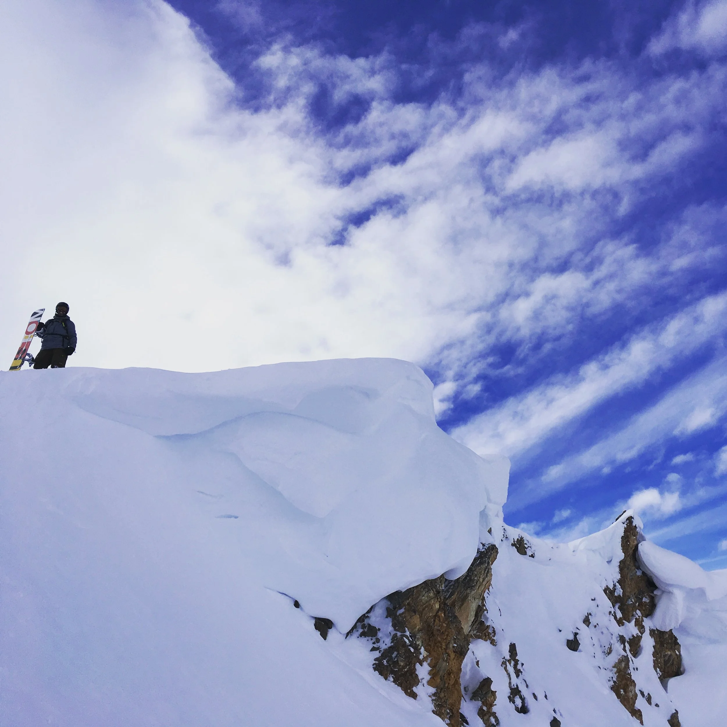 A person standing on a snowy mountain ridge holding a snowboard, looking at the snow-covered slope and partly cloudy sky.