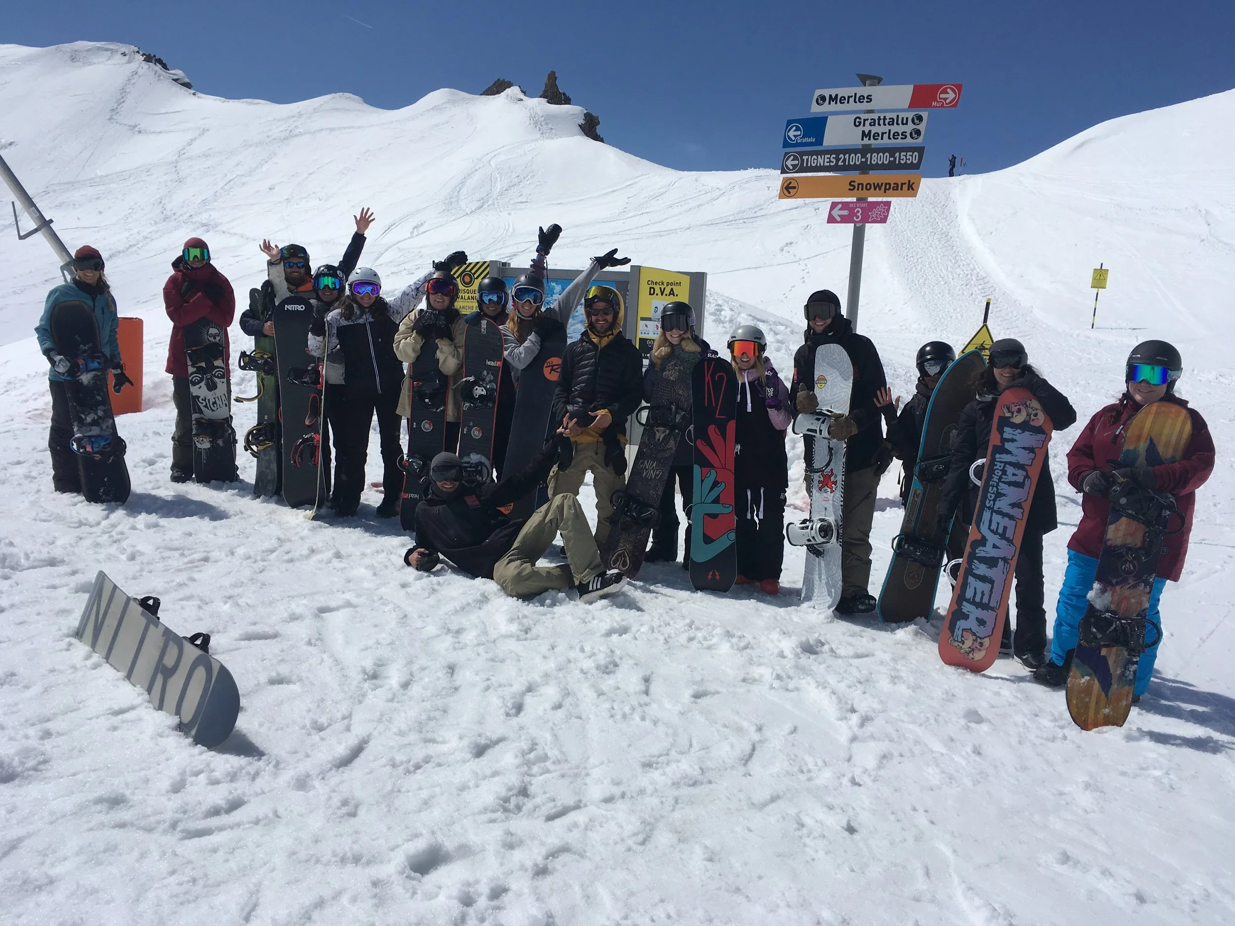A group of people in snowboarding gear standing on snow at a ski resort with snow-covered mountains in the background. They are holding snowboards and wearing helmets and goggles, with some waving or posing for the photo.