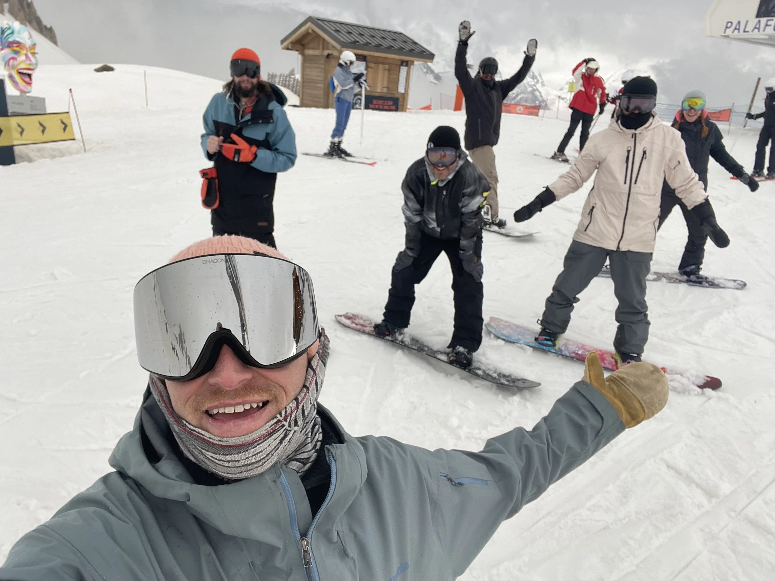 Group of people on a snowy mountain, some snowboarding, with one person in the foreground taking a selfie, smiling, wearing snow goggles and winter jacket.