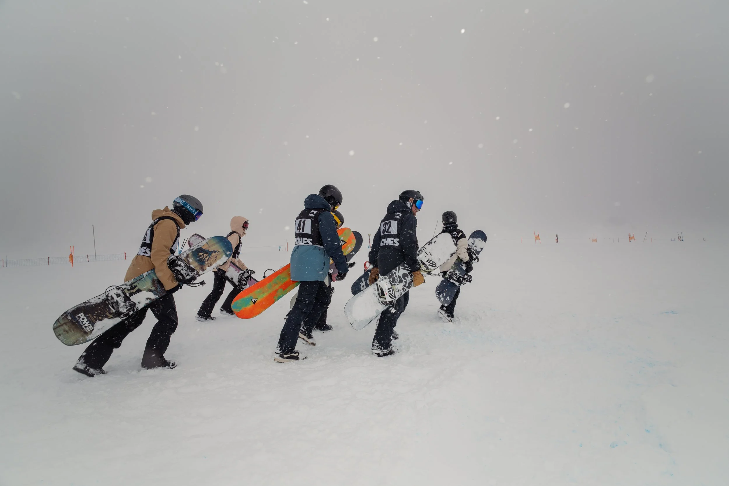 A group of six snowboarders walking through snow with their snowboards in hand on a foggy, snowy day, wearing helmets and winter gear.