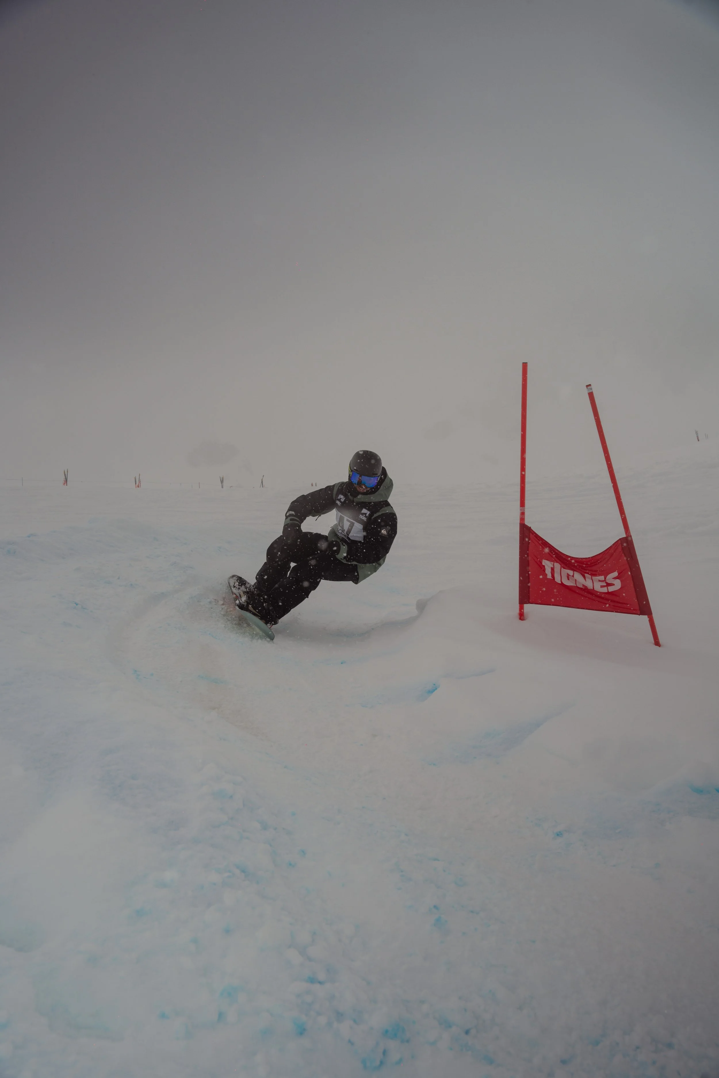 A snowboarder in black gear making a turn on a snowy slope near a red flag marked 'Tignes' in a foggy, overcast environment.