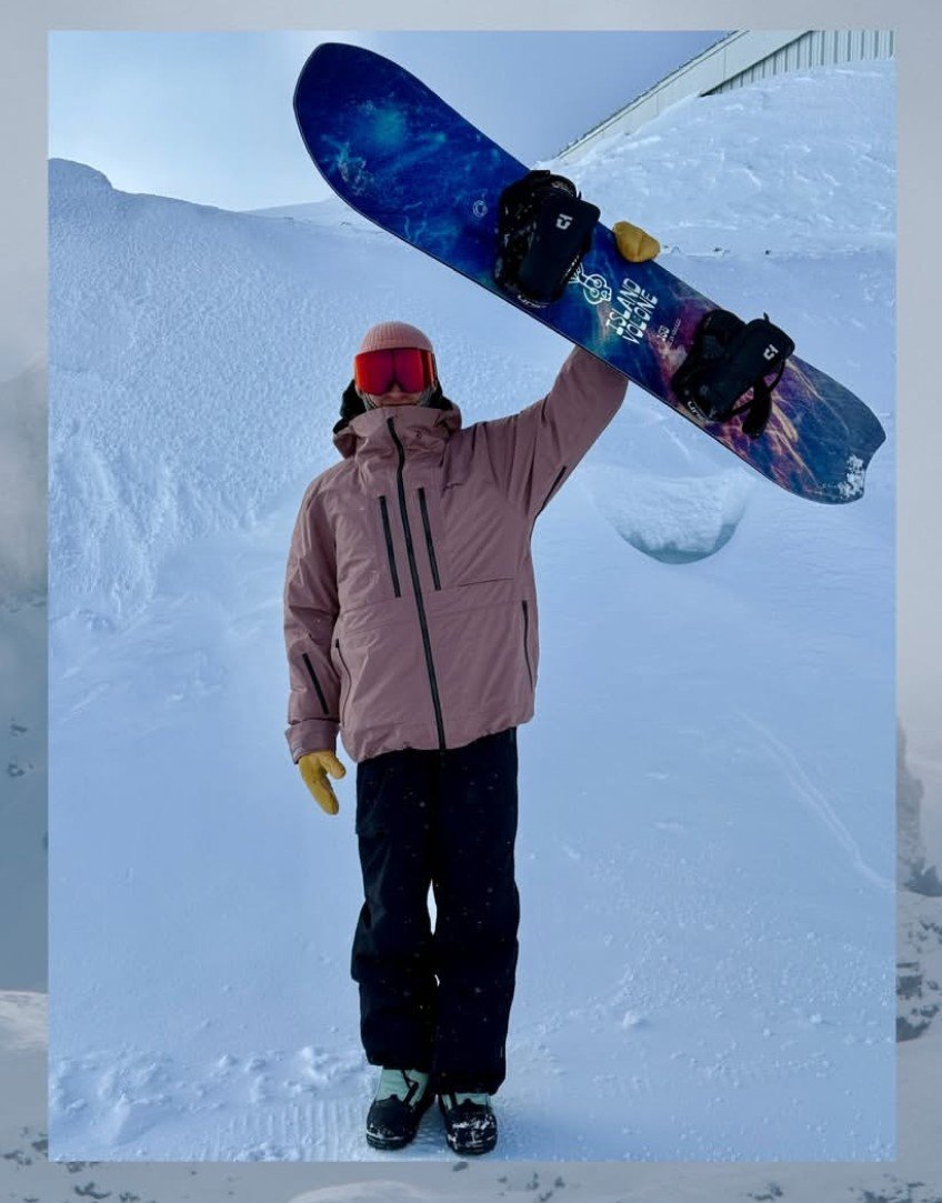 Person standing in snow holding a snowboard above their head, wearing a pink jacket, yellow gloves, a red helmet, and snowboarding boots, with snow-covered ground and a building in the background.