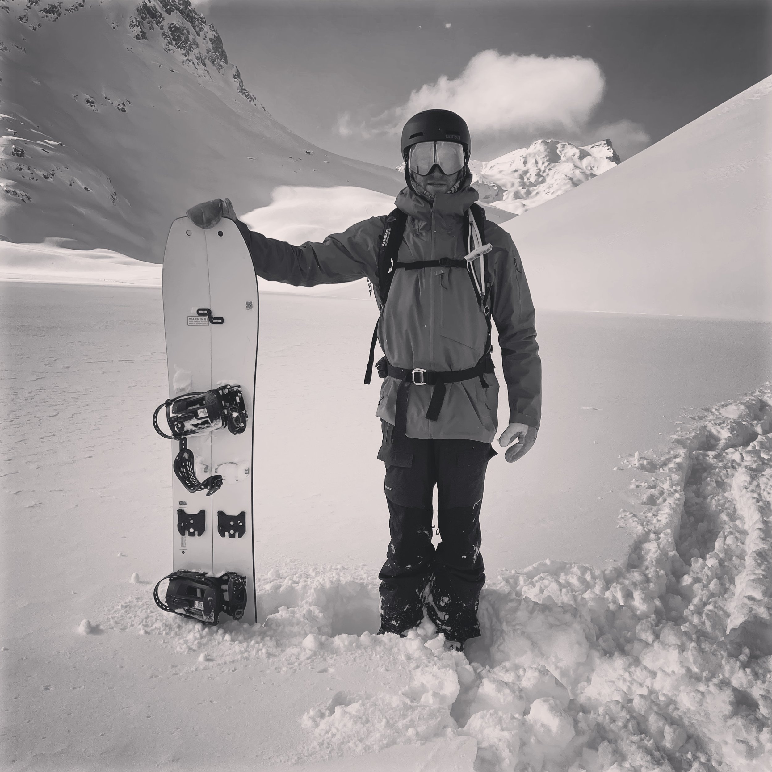A person dressed in winter gear, including a helmet and goggles, standing in snow with a snowboard in hand during a snowy mountain landscape.