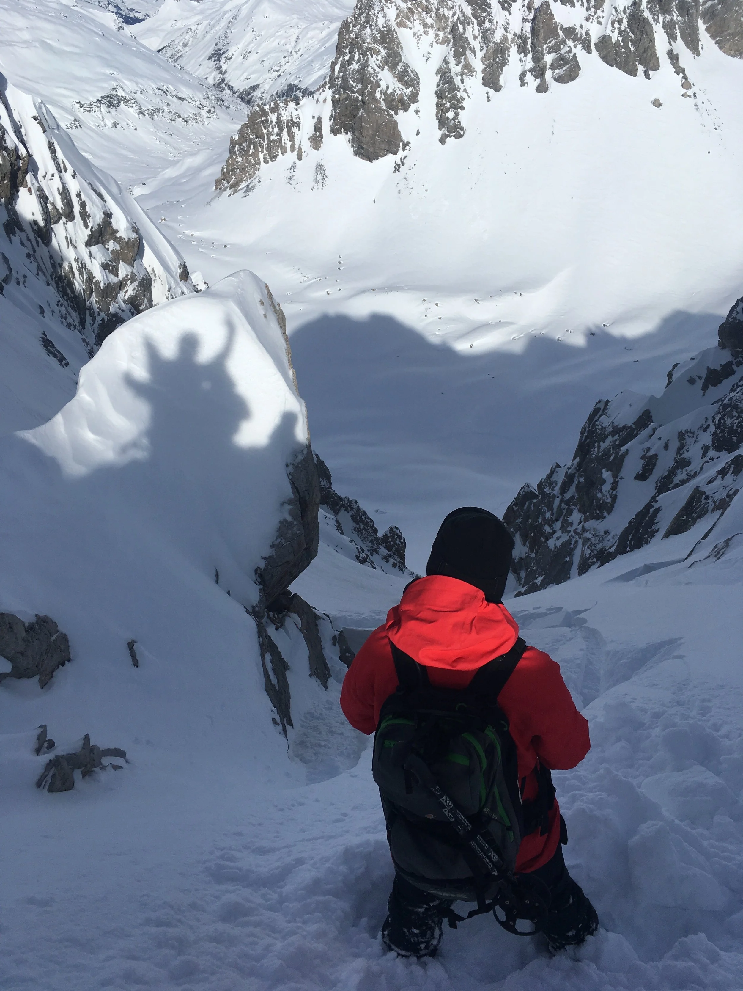 A person dressed in a red jacket and black pants kneeling in deep snow in a snowy mountain valley.
